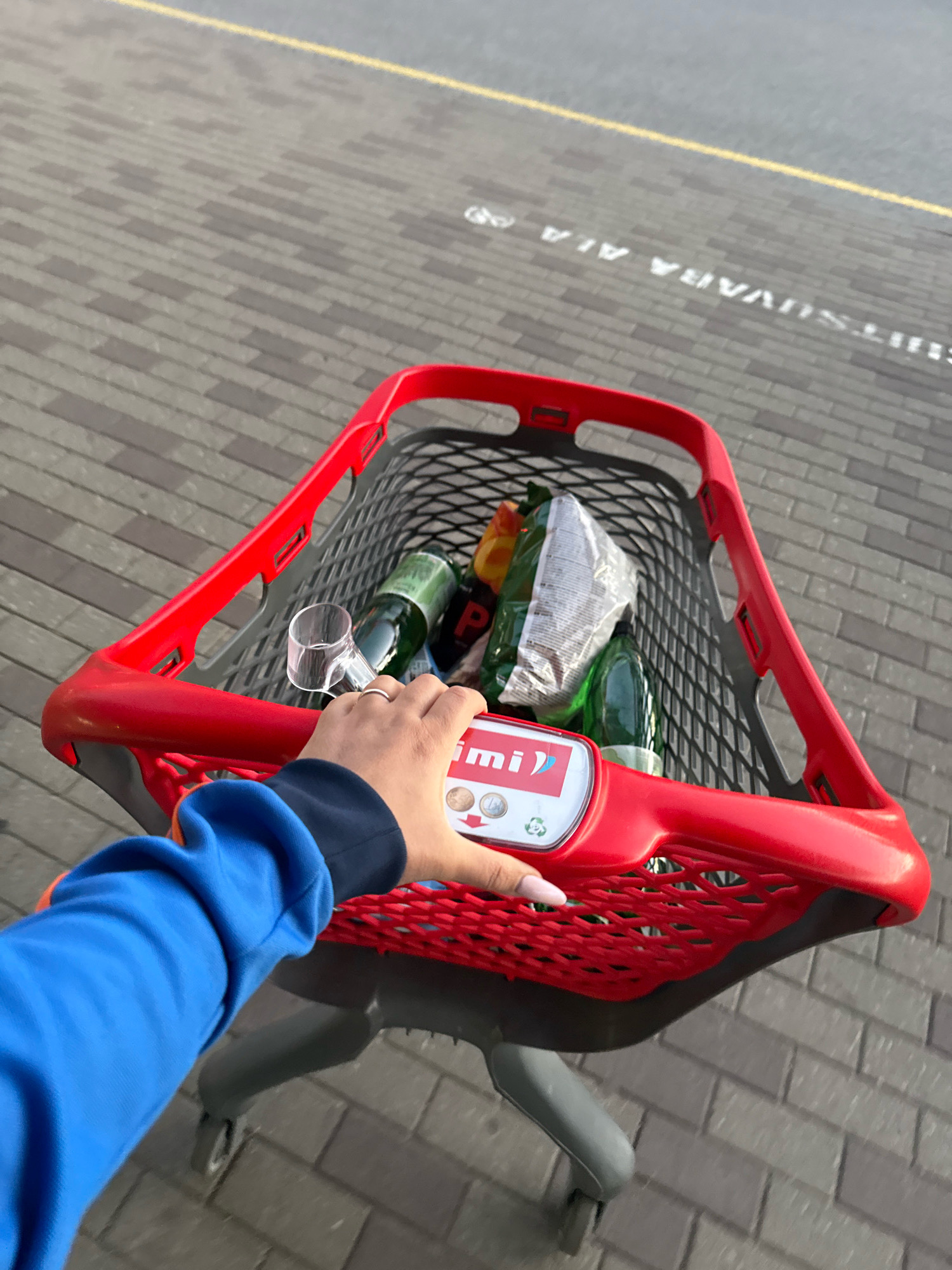 The image shows a first-person perspective of someone pushing a red shopping cart on a paved walkway with a yellow line marking. The shopping cart contains several green bottles, a bag of what appears to be snacks, and a small clear plastic item. The hand pushing the cart has a ring on one finger and long fingernails with a light pink polish. The person is wearing a bright blue long-sleeved shirt. In the background, white lettering is visible on the pavement, which reads "SIHTSUVARU ALA" and also text that appears to be "ALA" further down. This lettering is consistent with signage found in parking areas or designated zones in Estonia. The overall scene suggests an outdoor urban environment, likely a parking lot or an area associated with a shopping center or supermarket in Tallinn, Estonia.