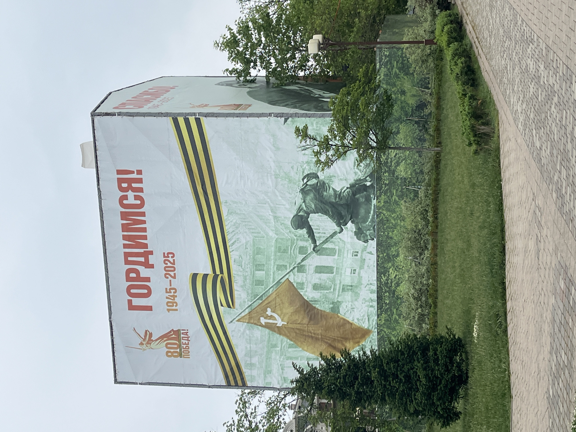 The image depicts a large banner in Krasnodar, Russia. The banner commemorates the 80th anniversary of victory in World War II, with the dates 1945-2025 displayed. The Russian text "ГОРДИМСЯ!" is prominently featured, meaning "We are proud!". The banner includes imagery associated with the Soviet Union, such as the Hammer and Sickle flag, the Ribbon of Saint George, and a sculpture of a soldier raising a flag. The banner is set against a backdrop of a park or green space, with trees and a paved walkway visible.