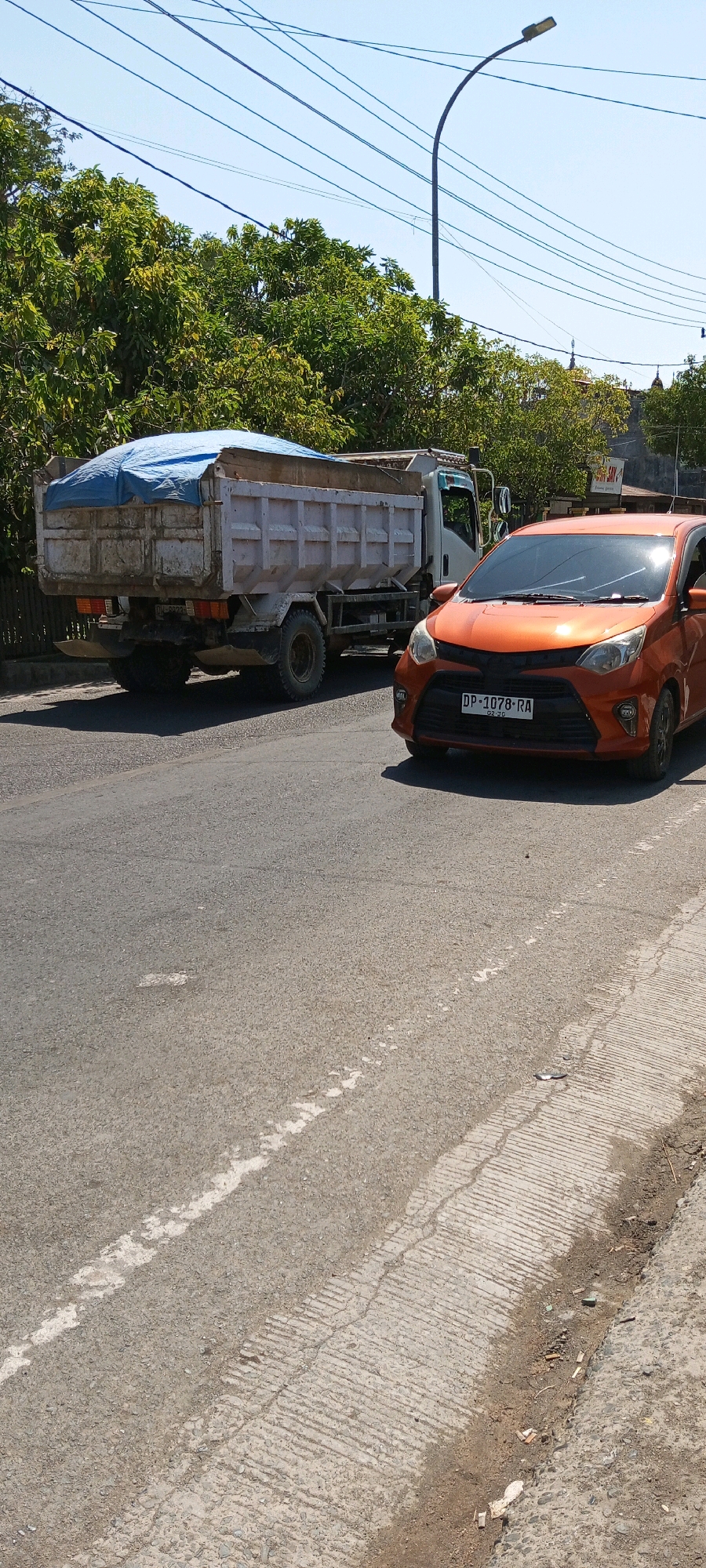 An orange car is parked on a paved road. The car is facing right and its license plate reads "DP 1078 RA". Behind the car is a white dump truck. The dump truck is covered with a blue tarp and its license plate is partially visible. The road is lined with trees and a sidewalk. The scene is illuminated by the sun, indicating it is daytime. The overall environment appears to be urban or suburban, possibly in Sengkang, Indonesia given the context.