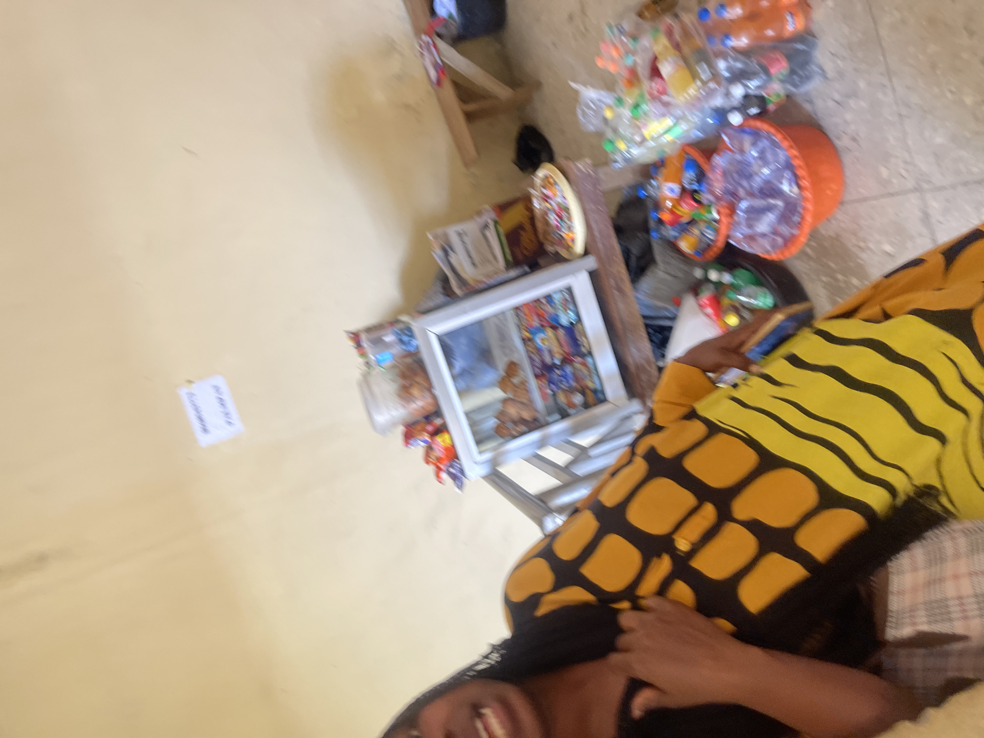 In Rafin Par, Nigeria, a woman wearing a yellow and black patterned dress stands near a display of various goods, including bottled drinks, snacks, and candies. A small refrigerated display case contains more snacks. A wooden bench is visible in the background. The scene appears to be an outdoor market stall or a small shop.