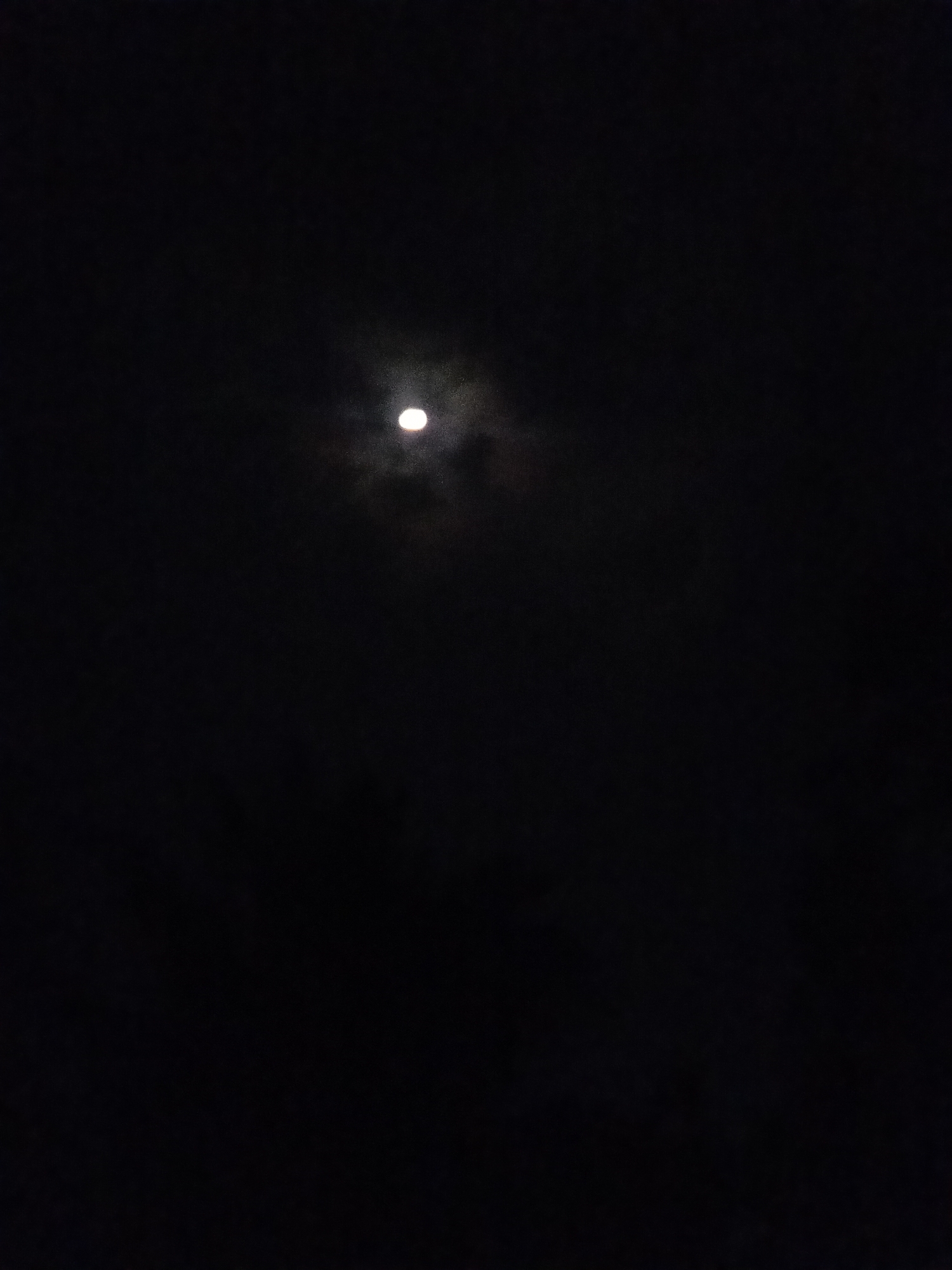 The image depicts a nighttime scene with a full moon slightly obscured by clouds. The moon is centrally located in the frame, emitting a soft glow against the dark sky. There are no visible people, objects, or buildings in the image, indicating an open sky environment. The location in Makale, Indonesia, is noted, but the image itself does not contain any specific visual cues to confirm this location. The overall impression is one of a serene, natural nighttime setting.