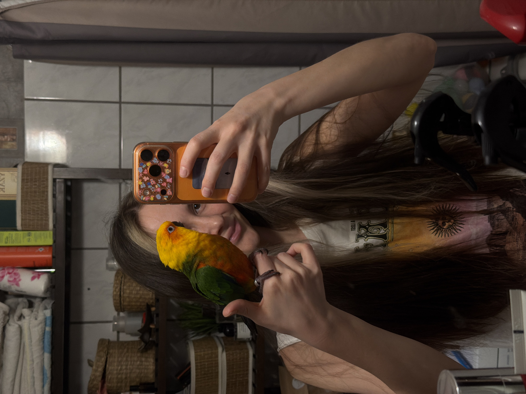 A person is holding a parrot while taking a selfie in a bathroom. The person is holding an orange smartphone with a colorful case. The parrot is perched on the person's hand. The bathroom has white tile walls and a shelf with books and other items. The location is Khimki, Russia.