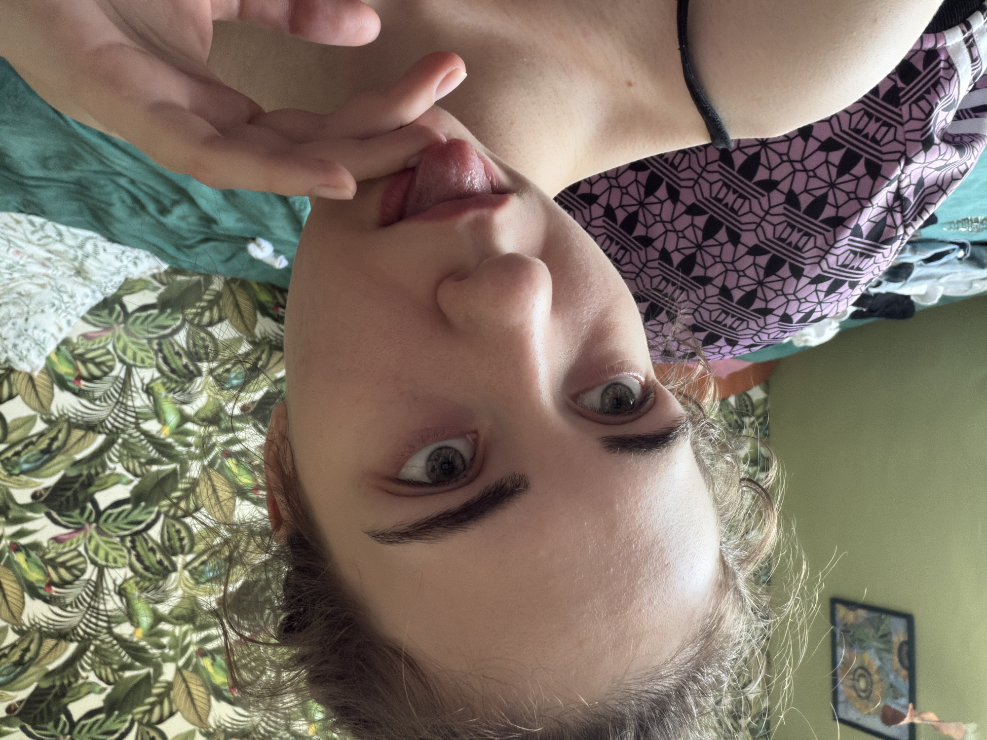 The image shows a close-up selfie of a young woman indoors. She has light skin, green eyes, and brown hair pulled back in a messy bun. Her tongue is sticking out and touching her finger. She is wearing a black spaghetti strap top. In the background, there is a wall with tropical-themed wallpaper featuring green leaves and birds. To the left, a framed picture of sunflowers hangs on a green wall. A bed with a teal-colored duvet and a patterned purple blanket is visible. The scene appears to be a bedroom. The location is not specified within Zavodoukovsk, Russia.