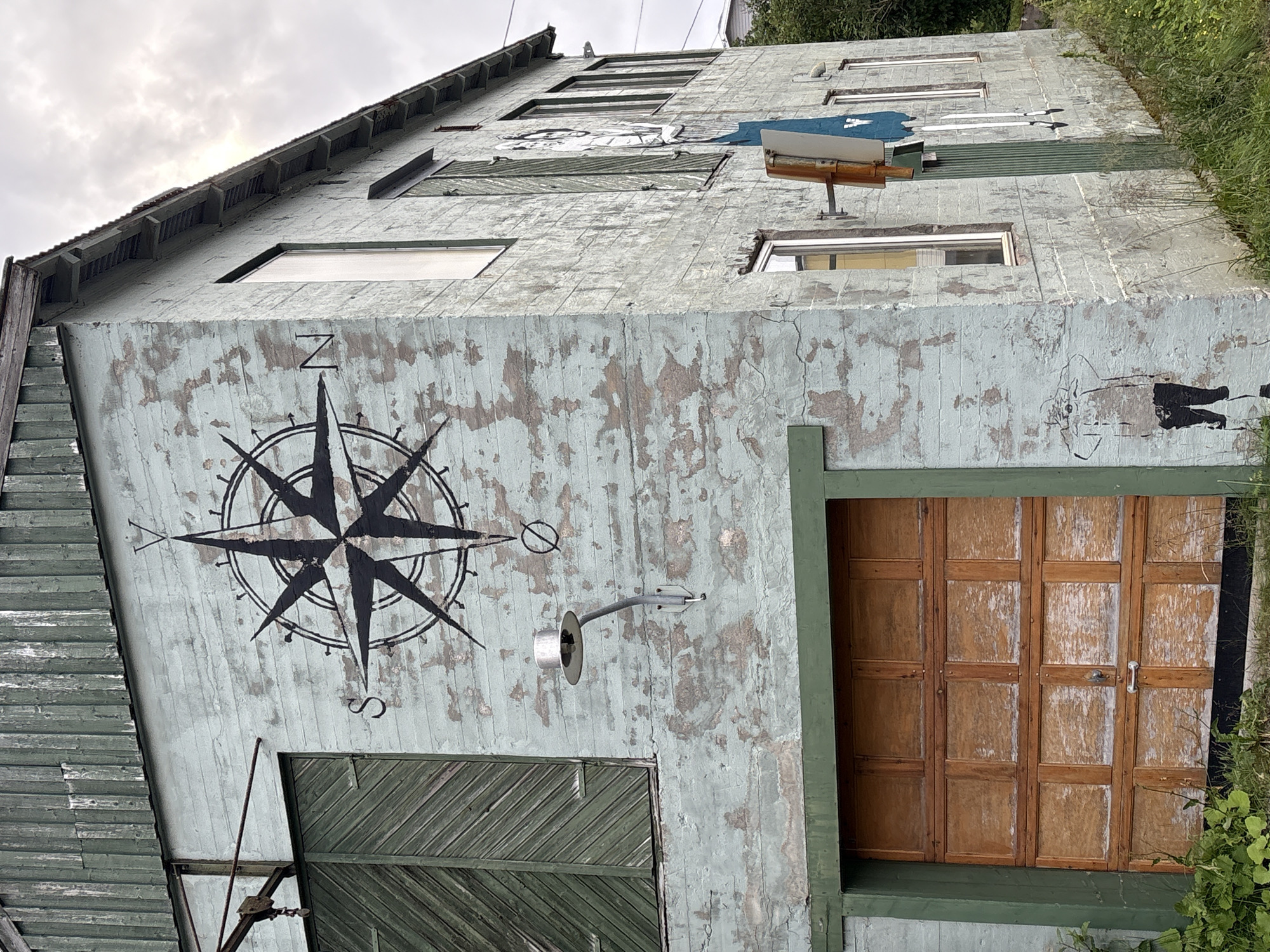 The image shows a two-story concrete building with peeling light blue paint. The building is located in Gryllefjorden, Norway. A large black compass rose is painted on the side of the building. A wooden garage door is visible on the ground floor, and a green paneled door is visible on the first floor. There are several windows on both floors. A street lamp is mounted on the wall. Graffiti of a person is visible on the lower part of the wall. The building is surrounded by grass and some foliage. The sky is overcast.