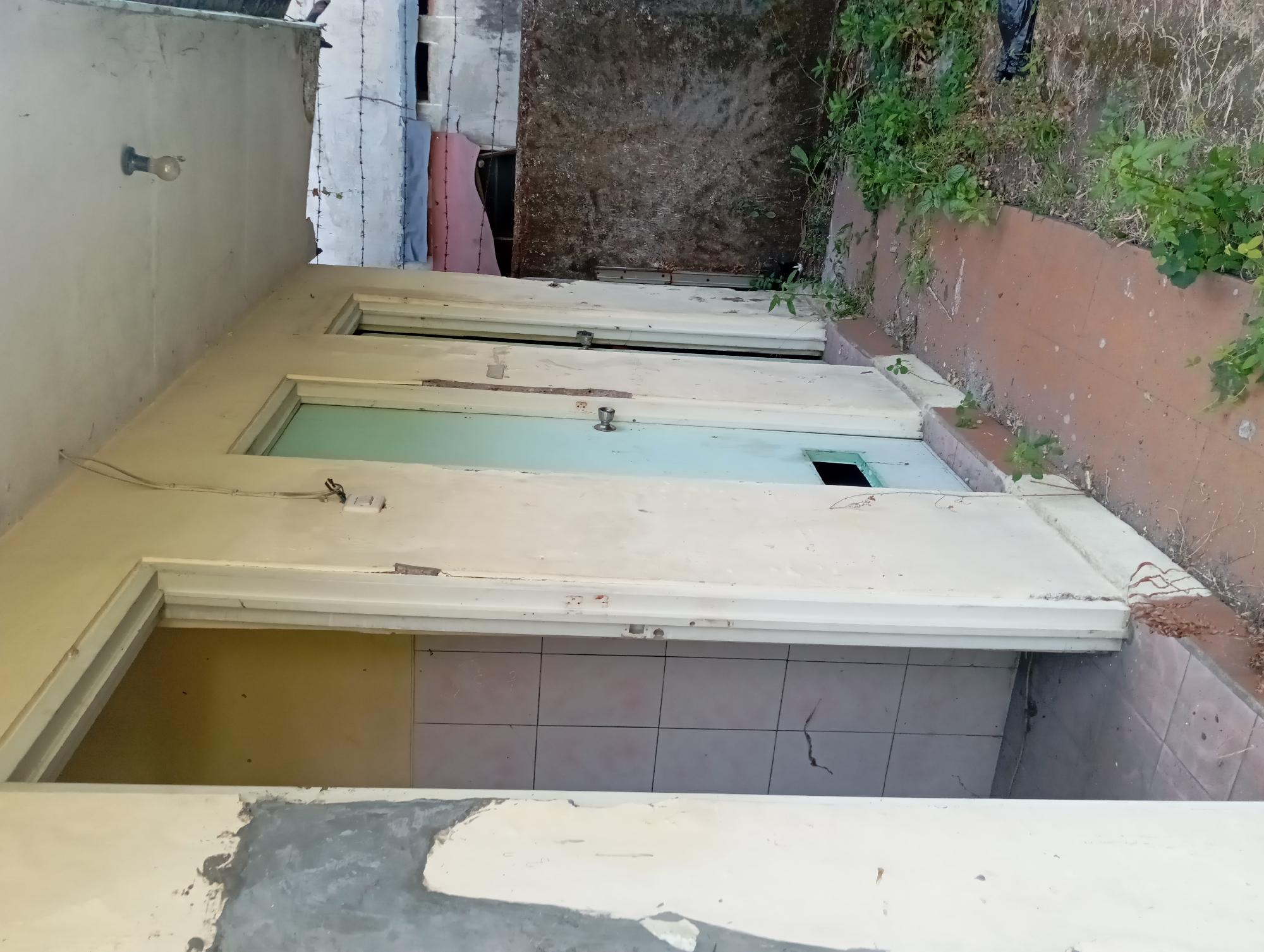 The image depicts the exterior of a dilapidated building with two doorways and an overhang. The walls are a pale yellow, showing signs of wear, peeling paint, and patches of unfinished concrete or plaster. The left doorway opens into a tiled room. The right doorway is partially open, revealing a light blue door with a silver doorknob and a rectangular opening near the bottom. A bare lightbulb hangs from the ceiling of the overhang. Beyond the building, a wall topped with barbed wire is visible, suggesting a security measure or a boundary. Overgrown vegetation and some debris, including a black plastic bag, are present on the ground in front of the building. The scene appears to be in a state of disrepair and neglect, consistent with an abandoned or uncared-for location in the city of Pandaan, Indonesia.