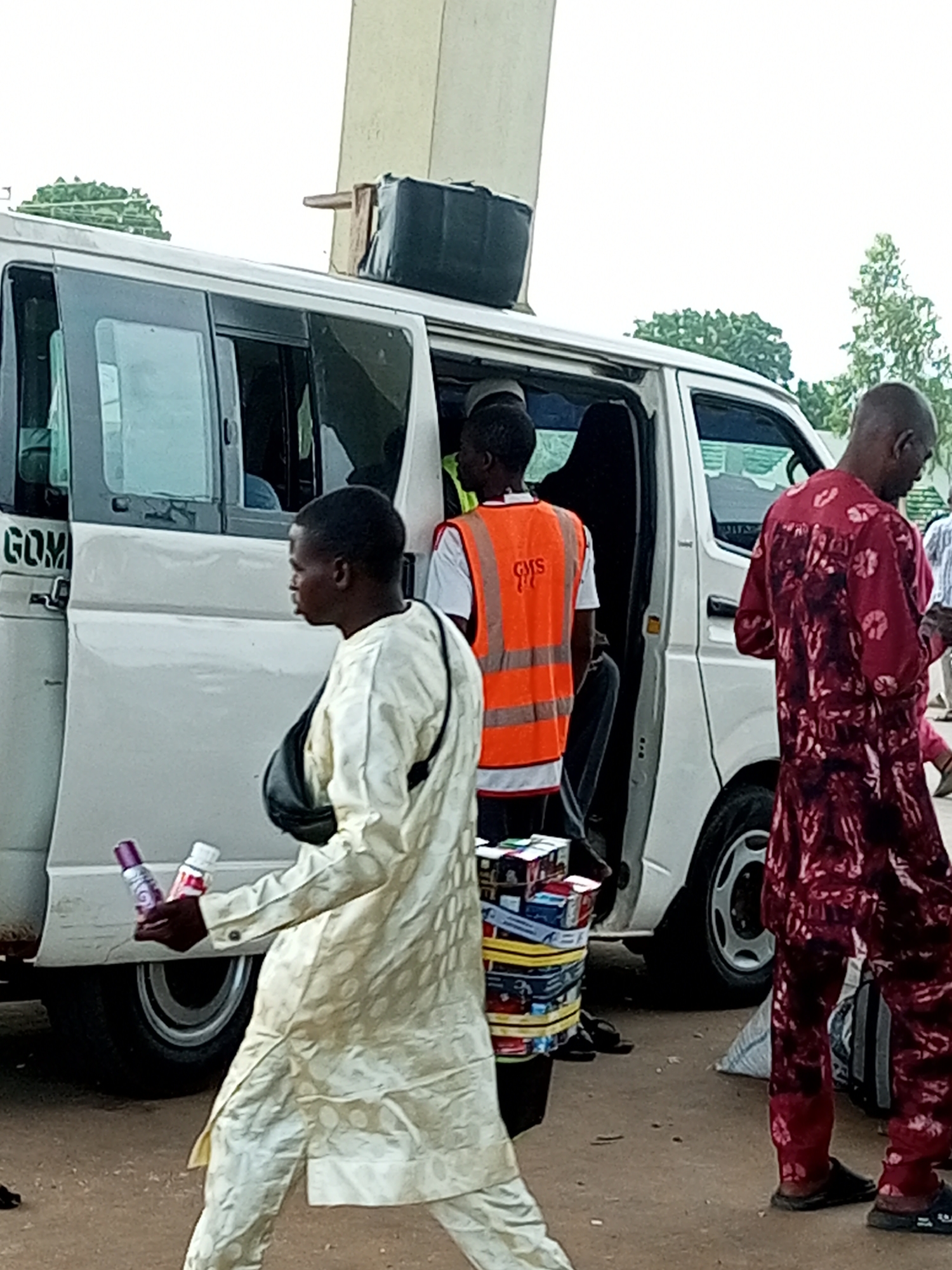 The image depicts a street scene in Gombe, Nigeria, with a white van as the central object. Several individuals are present around the van. In the foreground to the left, a person in a light-colored patterned outfit is holding two bottles, possibly air fresheners or spray cans. They are wearing a dark bag over their shoulder. To the right of the van, a person in a dark red patterned garment is also visible. Inside the van's open doorway, another person is wearing an orange safety vest over a white shirt, with the logo "GMS" visible on the vest. There are stacks of what appear to be small boxes or packages, possibly products for sale, tied together with yellow straps near the van's entrance.
The setting appears to be outdoors, with a sandy or dirt ground. Trees are visible in the background, suggesting a somewhat rural or semi-urban environment. The sky is overcast, indicating a cloudy day, and the lighting suggests it might be daytime, possibly late morning or afternoon. There are no clear indicators of specific emotions, but the general atmosphere seems to be one of commercial activity or transit. The text "GOM" is visible on the side of the white van, likely part of the vehicle's branding or destination.