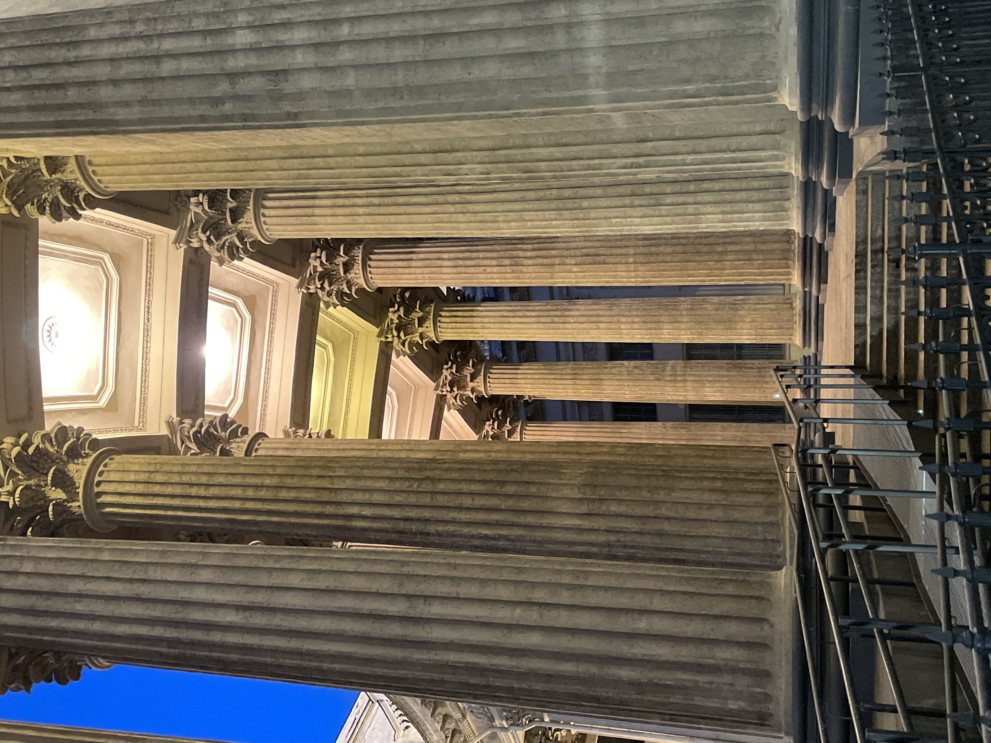 The image depicts a majestic architectural scene, likely the facade or portico of a grand classical building, viewed from a low angle looking upwards. Several colossal, fluted columns crafted from light-colored stone dominate the composition. These columns are adorned with intricately carved Corinthian capitals featuring detailed acanthus leaves and volutes, supporting a heavy entablature and ceiling.
Above the columns, the ceiling is visible, characterized by recessed rectangular panels, or coffers, which are outlined with decorative moldings. Within some of these panels, light sources emit a warm, golden glow, illuminating the architectural details. This artificial lighting, combined with the deep, dark blue sky visible through the columns on the left side of the frame, suggests the photograph was taken at night or during the deep twilight hours.
To the right, a dark, elaborate metal railing or gate is partially visible, hinting at an adjacent staircase or another structural element. The overall impression is one of monumental scale and historical grandeur, characteristic of the classical architecture found in cities like Saint Petersburg, Russia. There are no people, explicit activities, or visible text in the image.