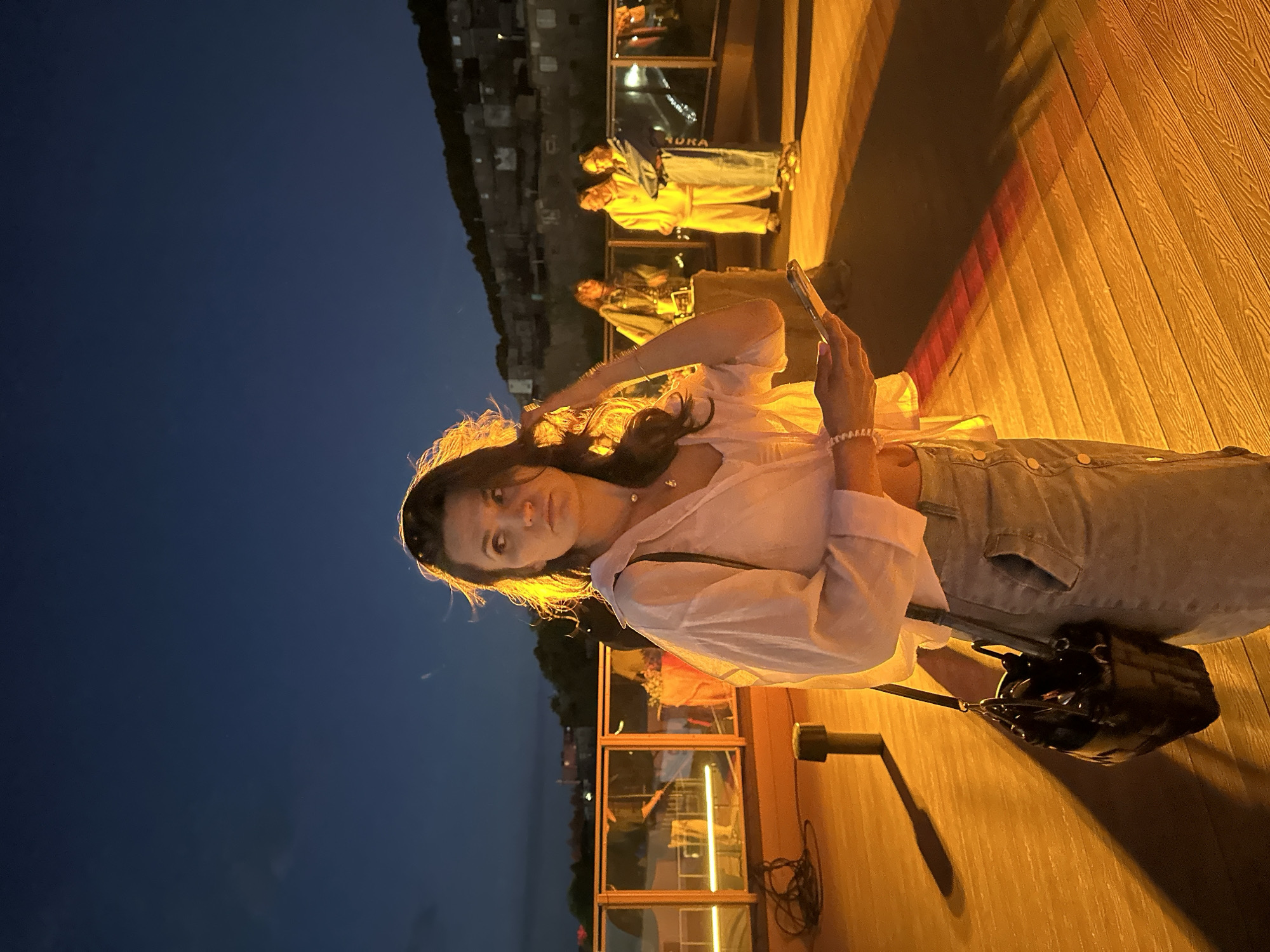 An outdoor nighttime scene shows a young woman in the foreground, standing on a wooden deck and looking to her right. She is wearing a light-colored open-collared shirt, a light-colored skirt, and a black purse hangs from her left shoulder. Her hair is dark and appears windswept. She holds a mobile phone in her right hand. In the background, several people are visible, standing near a railing. Behind them, a hillside settlement with lights is visible against a dark blue sky. The scene appears to be in Petropavlovsk-Kamchatsky, Russia, possibly on a pier or waterfront area given the lighting and the style of the buildings on the hill. The lighting is warm and artificial, casting long shadows on the deck.