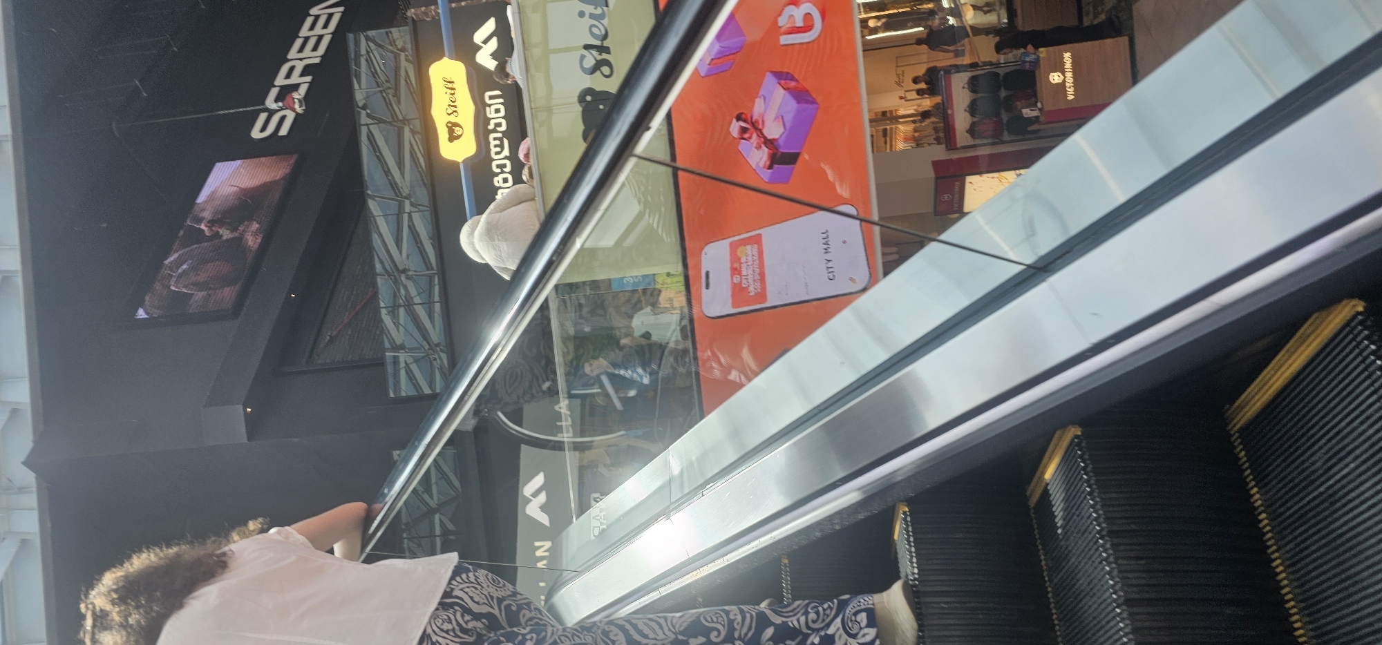 An overhead shot shows a person from behind ascending an escalator. The person has curly dark hair and is wearing a white shirt and patterned pants. The escalator has black treads with yellow indicator lines. The surrounding environment is a shopping mall with large screens displaying advertisements. One screen shows the word "SCREEN" in large white letters. Another screen displays a phone interface with the text "CITY MALL" and images of gift boxes. Further down the escalator, a store display for "Victorinox" is visible. In the background, other shoppers are present, and merchandise is displayed on shelves. The overall scene suggests a bustling commercial space.