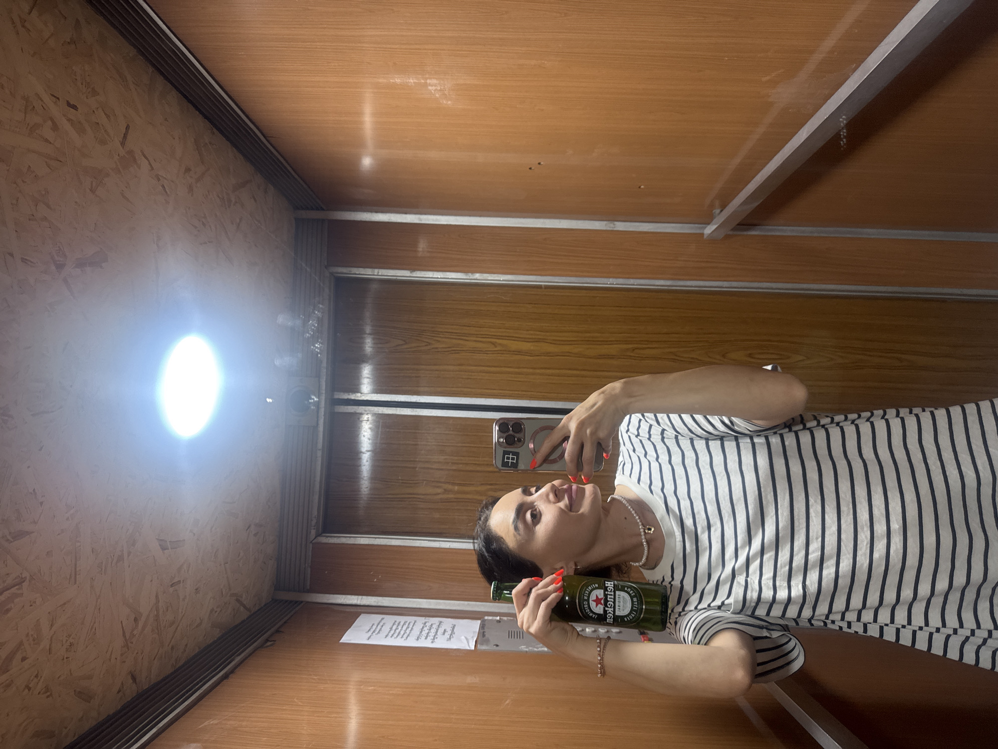 The image depicts a woman holding a bottle of Heineken beer and a smartphone, taking a selfie in what appears to be an elevator with wood-paneled walls and a wooden ceiling. The woman is wearing a horizontally striped t-shirt and a pearl necklace. Her fingernails are painted bright red. The elevator is lit by a bright overhead light. The walls are a warm brown color with a wood grain pattern. There is a white paper tacked to the left wall. The elevator doors are visible in the background. The scene is likely an interior residential or commercial building.
