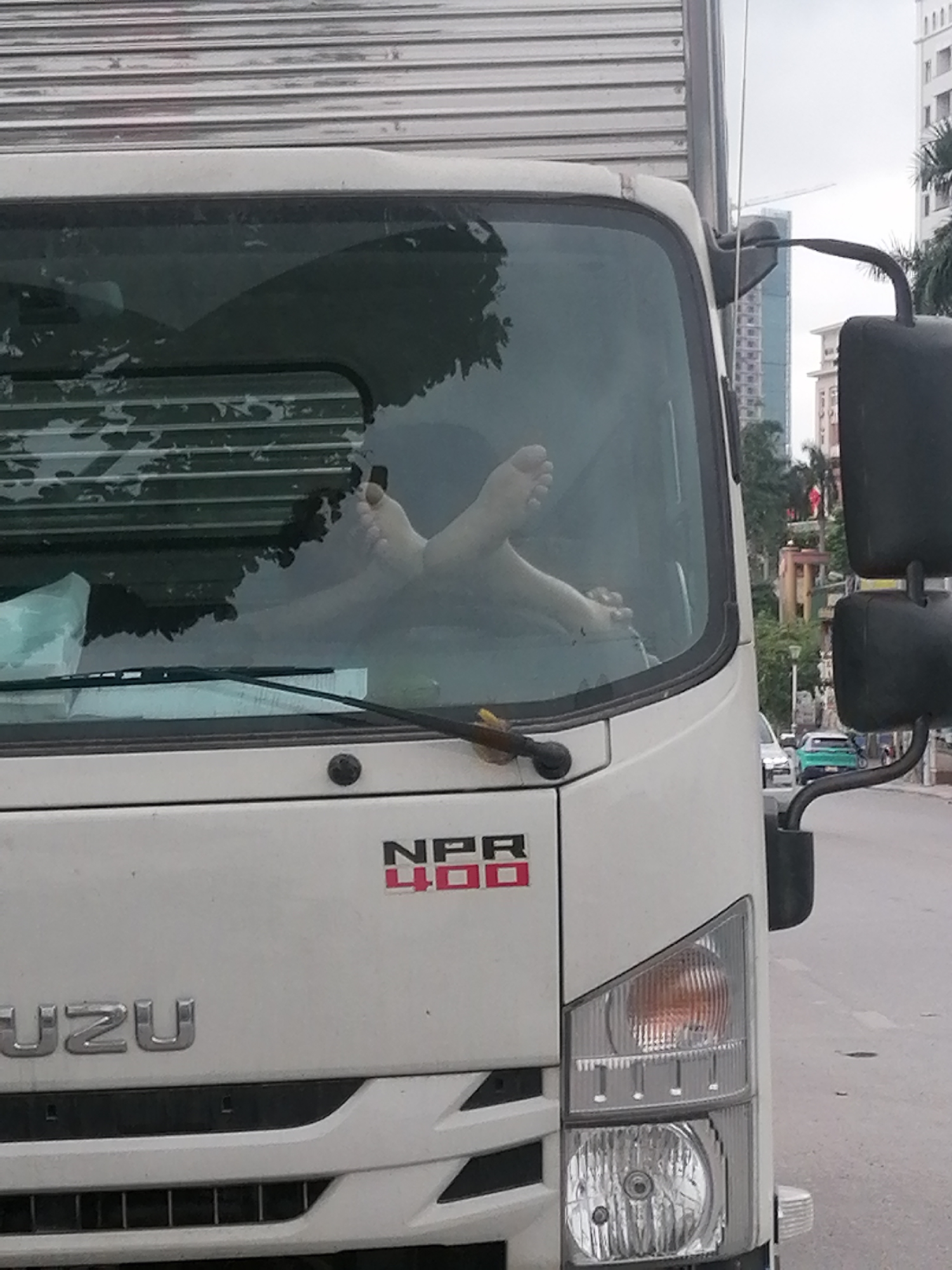 The image shows the front of a white Isuzu NPR 400 truck, parked or stationary on what appears to be a street in Bai Chay Ward, Vietnam. A notable detail is a pair of bare feet resting inside the truck's windshield, propped up on the dashboard or a similar surface, indicating a person is relaxing within the vehicle. The rest of the person's body is obscured by the dark interior of the cabin.
The truck features the "ISUZU" brand name prominently on its grille and "NPR 400" in red and black lettering on the front panel below the windshield. The vehicle has a large windshield, two wipers, and a substantial side mirror visible on the right. The corrugated metal side of the truck's cargo area is visible above the cab.
The background reveals an urban or suburban setting under an overcast sky. Multi-story buildings, including one with a visible construction crane, and green trees line the street. Other vehicles are faintly visible further down the road. The scene suggests a moment of rest during the daytime in a bustling environment.