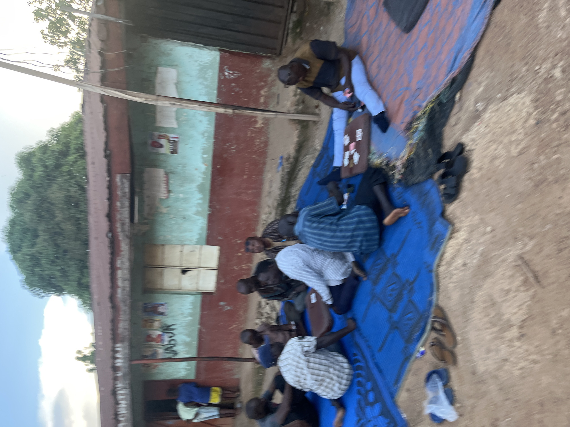 The image depicts a group of men resting on blue and red mats outdoors, likely in a communal setting in Jos, Nigeria. The scene appears to be during the daytime, with soft natural light suggesting it could be early morning or late afternoon. The men are dressed in casual clothing, including shirts and trousers, and some are barefoot while others wear sandals.
Several individuals are lying down or sitting, some appear to be dozing or resting, while others are engaged in quiet conversation or contemplation. One man in the foreground is wearing a checkered shirt and is seated with his legs crossed, looking towards the camera. Another man, in a white and blue striped shirt, is also seated. In the background, a building with a red and faded green facade is visible, featuring a window and what appears to be some posters or artwork on the wall. The ground is a mix of dirt and gravel, typical of an outdoor area in a developing region.
A few objects are scattered around the mats, including a brown bag or briefcase near one of the seated men, and a pair of brown sandals and a blue plastic bag on the ground to the right. The overall atmosphere seems relaxed and informal, suggesting a moment of pause or downtime for the individuals. There are no overt signs of distress or urgency, implying a peaceful or routine gathering. The vegetation in the background, with lush green trees, suggests a warm climate. The text "VASUR" is visible on the building wall, though its meaning or relevance is unclear without further context.