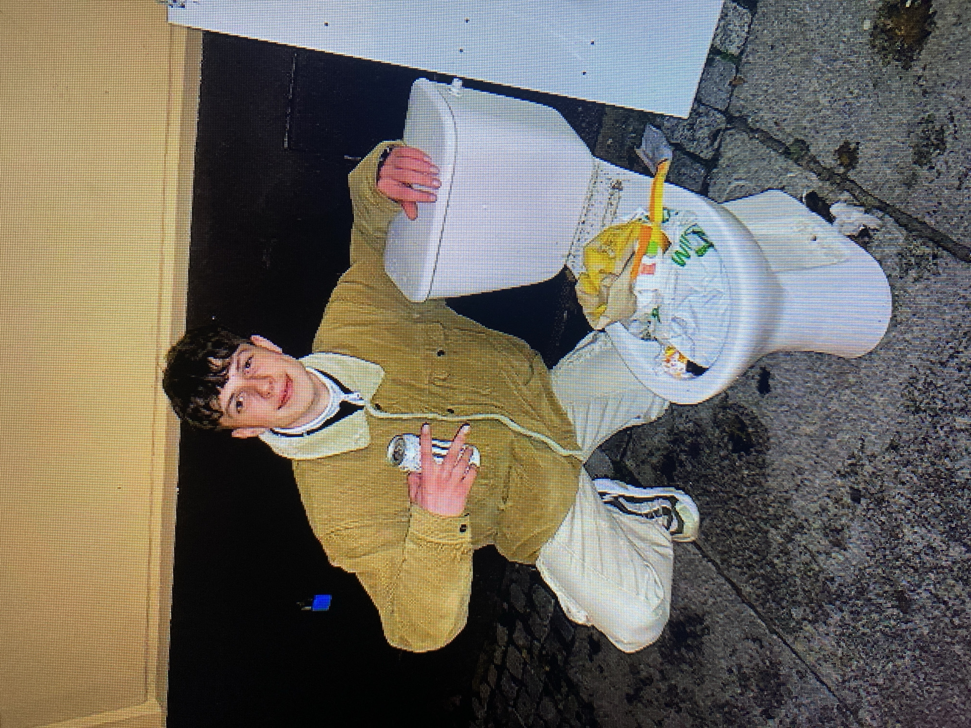 A young man with curly brown hair is kneeling on a cobblestone street next to a white toilet filled with trash. He is wearing a tan jacket over a white turtleneck and light-colored pants. He holds a can of beer in his right hand and rests his left hand on the toilet tank. The toilet is located outdoors, with a dark wall behind it and a light-colored vertical object to the right. The scene appears to be in the city of Poznan, Poland.