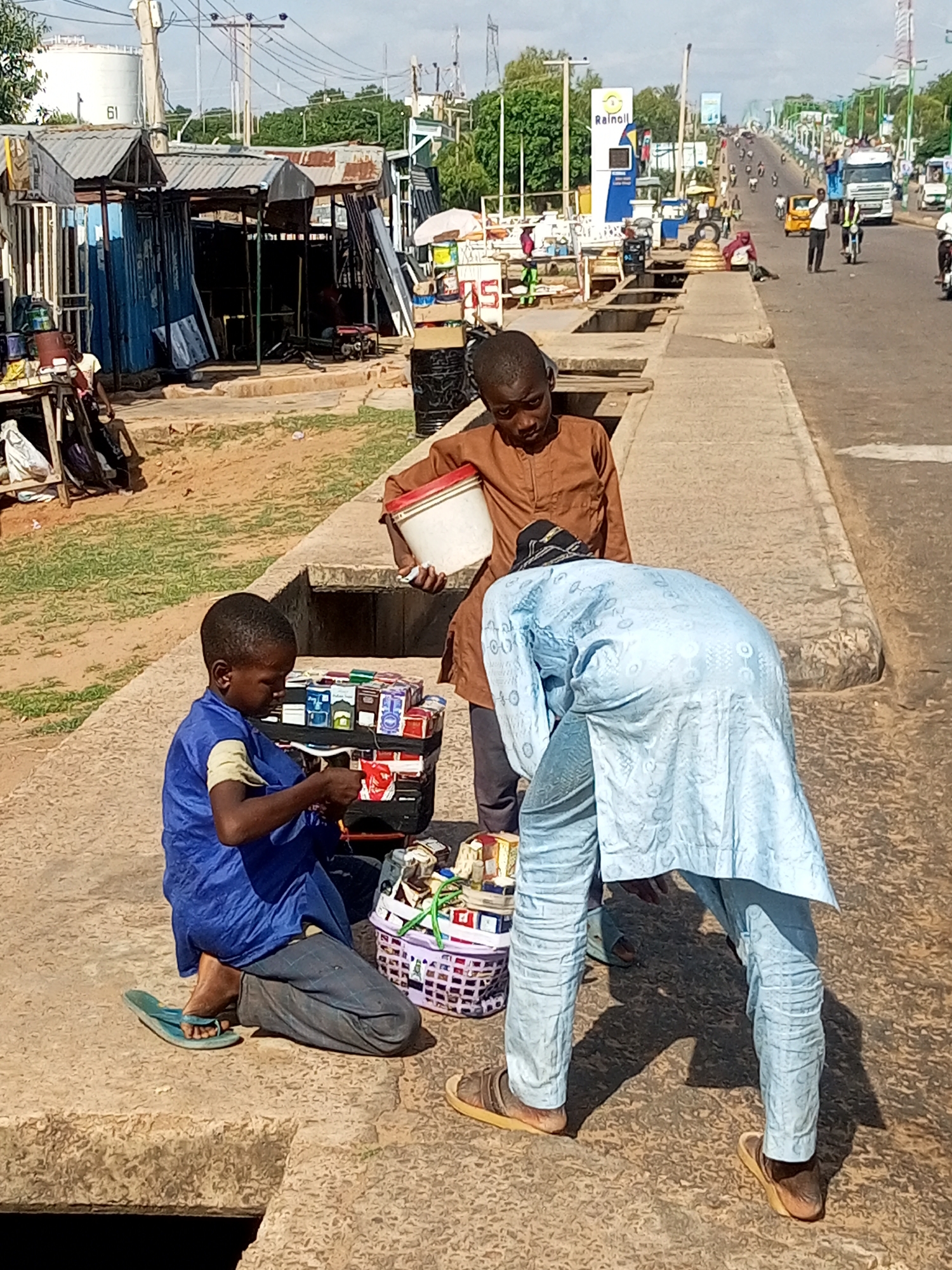 Three young men are engaged in selling goods on a sunny day in Tempure, Nigeria. The scene takes place on a paved area beside a busy road with traffic, including cars, trucks, and motorcycles. In the background, there are market stalls with corrugated metal roofs, a white fuel tank with the number "61" on it, and a prominent "Rainoil" sign.
The central subject is a boy in a blue tunic and grey trousers, sitting on a raised concrete edge and attending to a display of goods, possibly cigarettes or small packaged items. He wears flip-flops. Beside him, another boy in a brown tunic is holding a white bucket with a red rim. A third person, wearing a light blue patterned robe and trousers, is bent over, seemingly interacting with the goods or assisting in the sale. This individual also wears sandals.
The activity appears to be a street vending operation. The items for sale are displayed in baskets and on a makeshift stand, suggesting a lively informal economy. The sunlight suggests it is daytime, likely mid-morning or afternoon, given the strong shadows. The overall impression is one of active commerce in a bustling urban environment. No specific emotions are discernible from the subjects' expressions, but their engagement with the goods indicates a focus on their task.