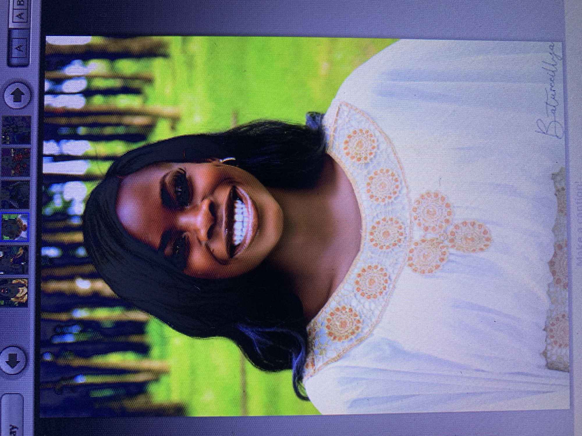 This is a close-up, vertically oriented photograph of a young Black woman. She is smiling warmly and looking directly at the camera. Her hair is dark and styled loosely around her face, with some strands falling over her shoulders. She is wearing a white garment adorned with an intricate embroidered pattern around the neckline and chest area. The embroidery features circular motifs in orange and gold tones.
The background is blurred but appears to be a lush green outdoor setting with vertical lines that suggest trees or foliage. The lighting suggests it might be daytime, with a soft, natural feel. The overall emotion conveyed is joy and confidence.
On the right side of the image, there is handwritten text in a script font, partially visible. It appears to read "Baturudiya".
The photograph is displayed on a screen, as indicated by the user interface elements visible on the left and top edges of the image. These include icons for navigation and text labels. The "ay" is visible at the bottom left. The context of Jos, Nigeria, provides a geographical reference for the image.
