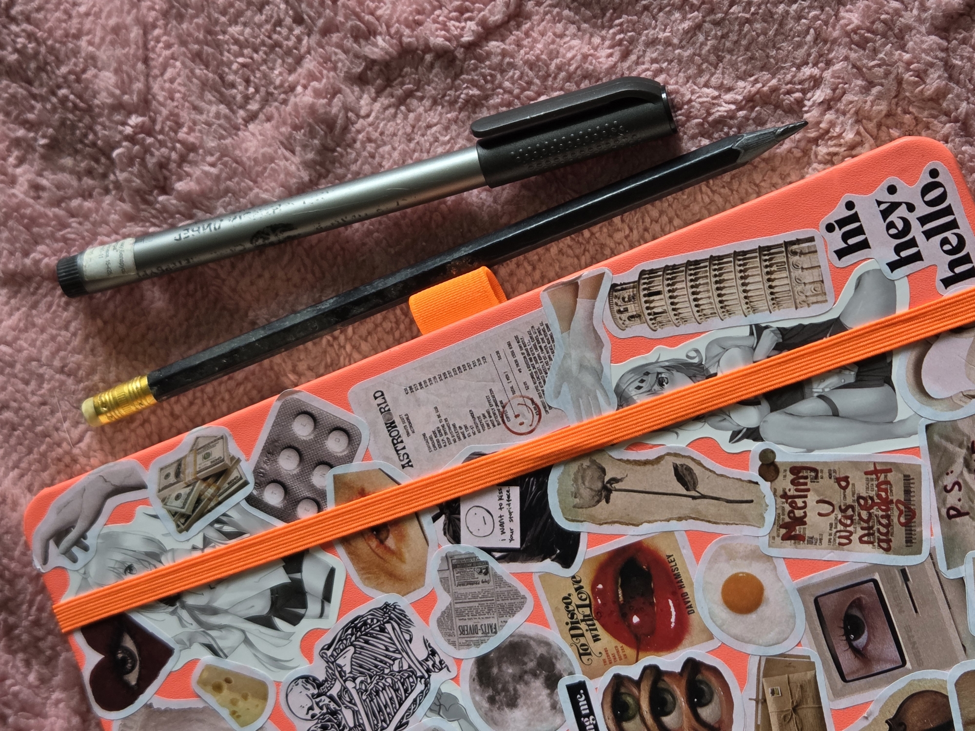 The image shows a coral-colored notebook covered in stickers, including images of the Leaning Tower of Pisa, eyes, faces, skeletons, money, pills, and text clippings. The notebook is secured with an orange elastic band. A silver pen and a black pencil lie on top of the notebook, against a pink, textured background.