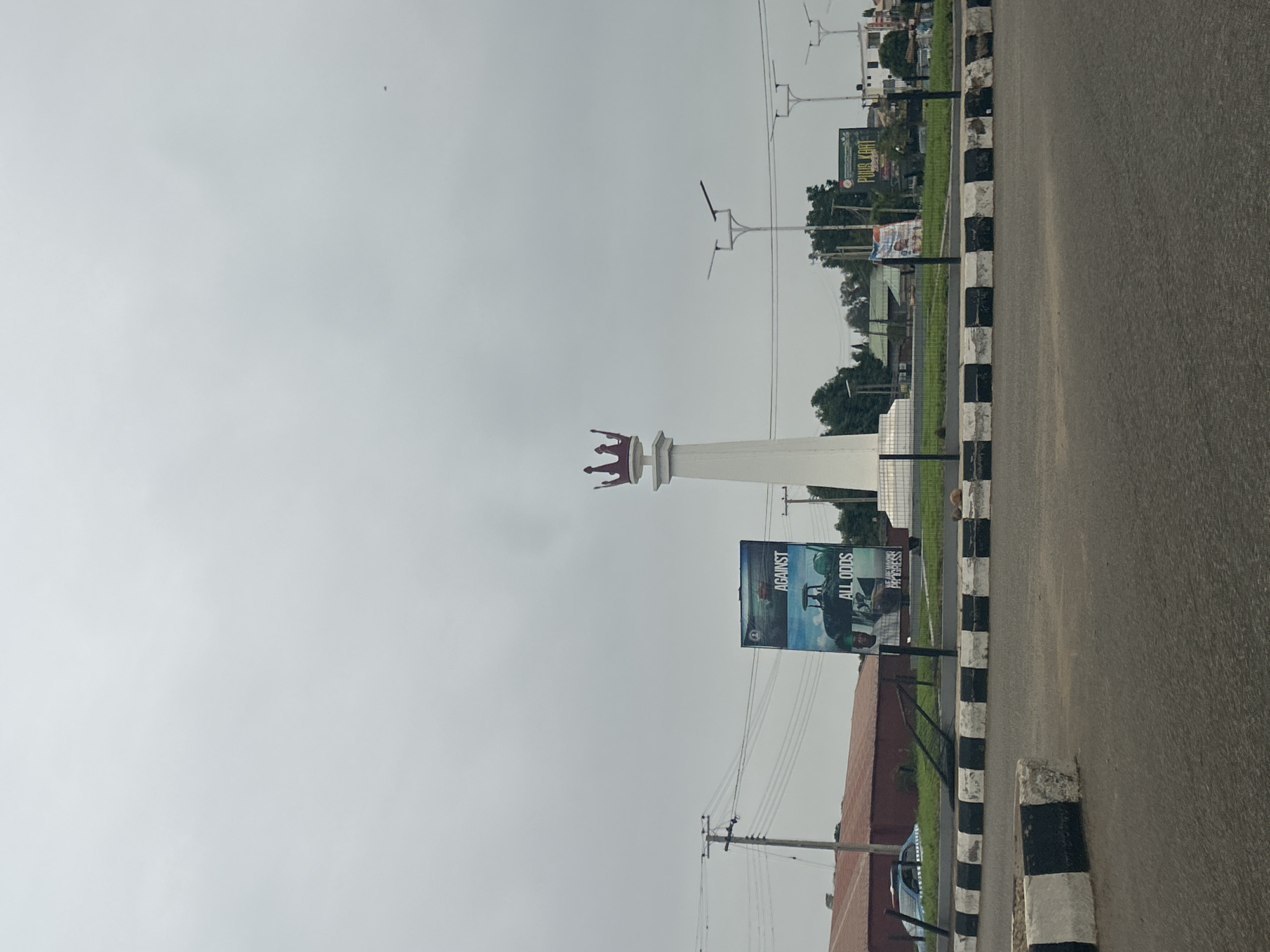 The image shows a street scene in Jos, Nigeria, featuring a monument topped with a red crown. The monument is white and stands near a roadside with a curb painted in alternating black and white stripes. Adjacent to the monument is a billboard advertising "Against All Odds". Overhead are power lines and several streetlights. In the background, there is greenery and a sign partially visible that reads "PLUS KART". The sky is overcast. A road is on the right.