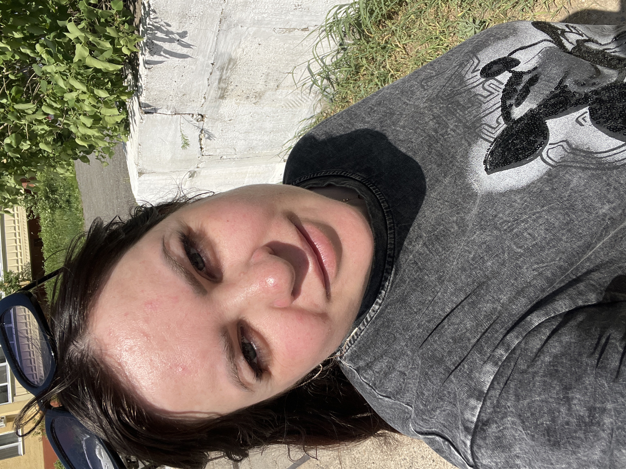 A woman with dark hair and fair skin is pictured outdoors, likely in a city environment. She is wearing a grey acid-wash top with a black and white design. Her sunglasses are visible in the lower left corner. The background includes a concrete wall, green foliage, and a building with multiple windows. The location is not discernible from the image; thus, the claim of being in the city of Noviy, Russia, cannot be confirmed.