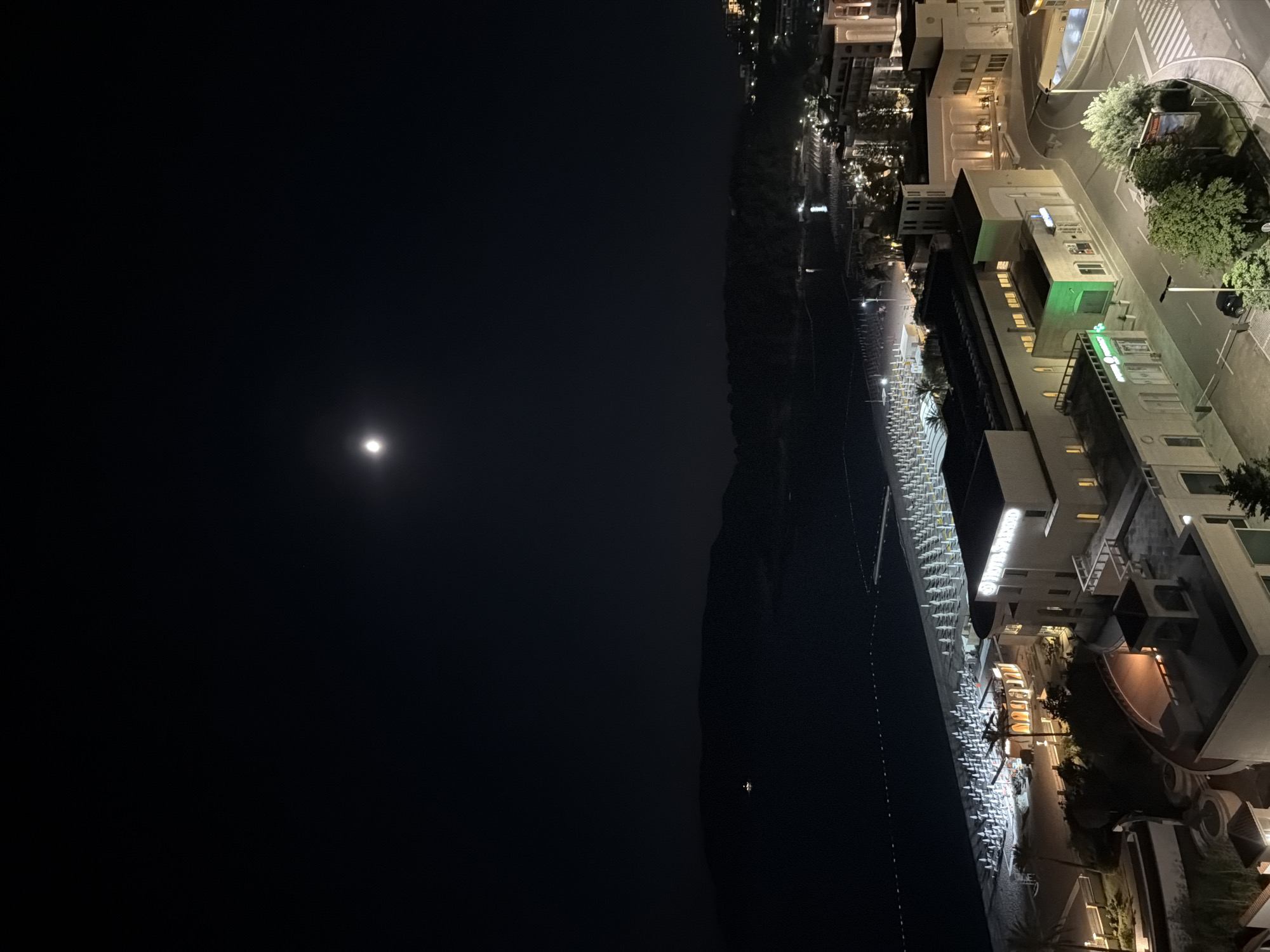 Nighttime view of Boreti, Montenegro. The moon is in the upper left quadrant. A dark mountain range separates the sky from the coastline. The coastline is illuminated with artificial light, revealing buildings and a beach with chairs. A brightly lit road with vegetation runs along the buildings.