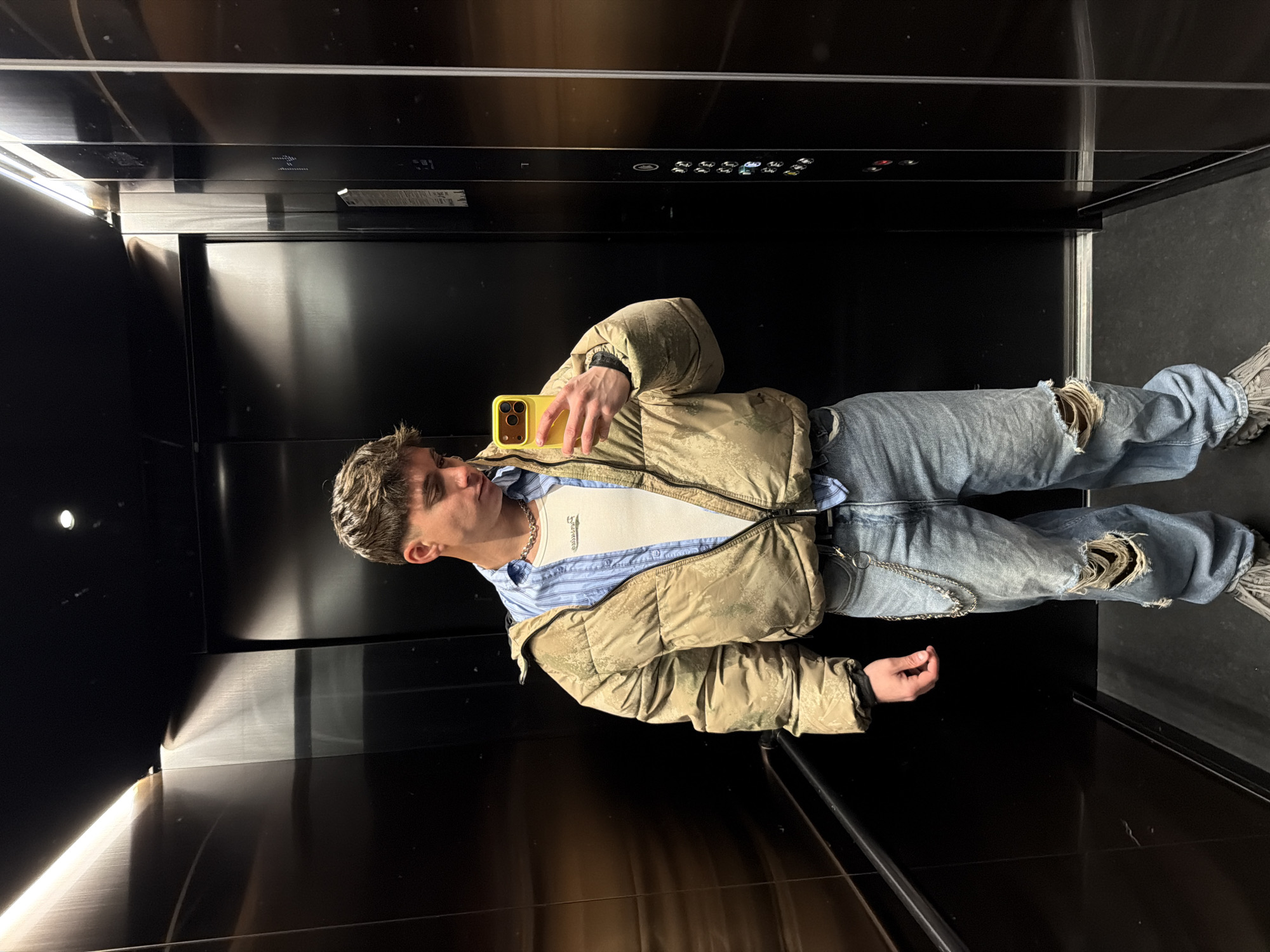 A young man is taking a selfie inside an elevator with black walls and ceiling. He is wearing a light brown puffer jacket over a blue and white striped shirt and a white undershirt. His light blue jeans are ripped at the knees, and he wears grey sneakers. He is holding a yellow smartphone in his right hand, angled to capture his reflection in the elevator's mirrored surface. The elevator's control panel is visible at the top of the frame. There is no specific information from the image that this is located in Sofia, Bulgaria.