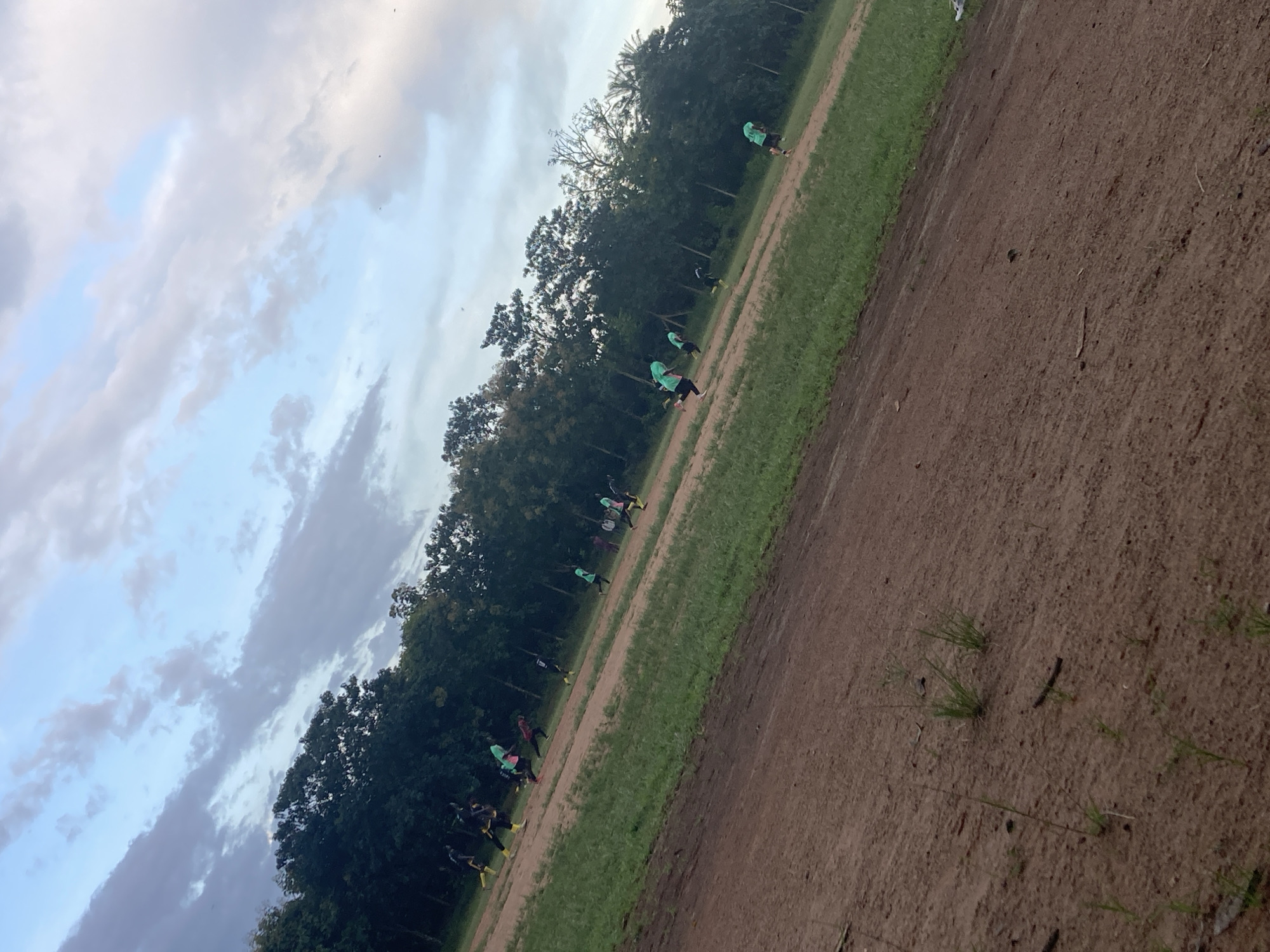 The image depicts a group of individuals, likely young men, engaged in athletic activity on a dirt field with patches of grass. They are dispersed across the field, some running and others appearing to be in motion. The participants are wearing athletic attire, with some distinguished by brightly colored shirts. The background consists of a dense line of trees, suggesting a rural or semi-rural setting. Above, the sky is partially cloudy, with a blend of blue and muted gray tones. The terrain is uneven and appears to be a natural or undeveloped sports ground. Based on the context, this scene is located in Miango, Nigeria.