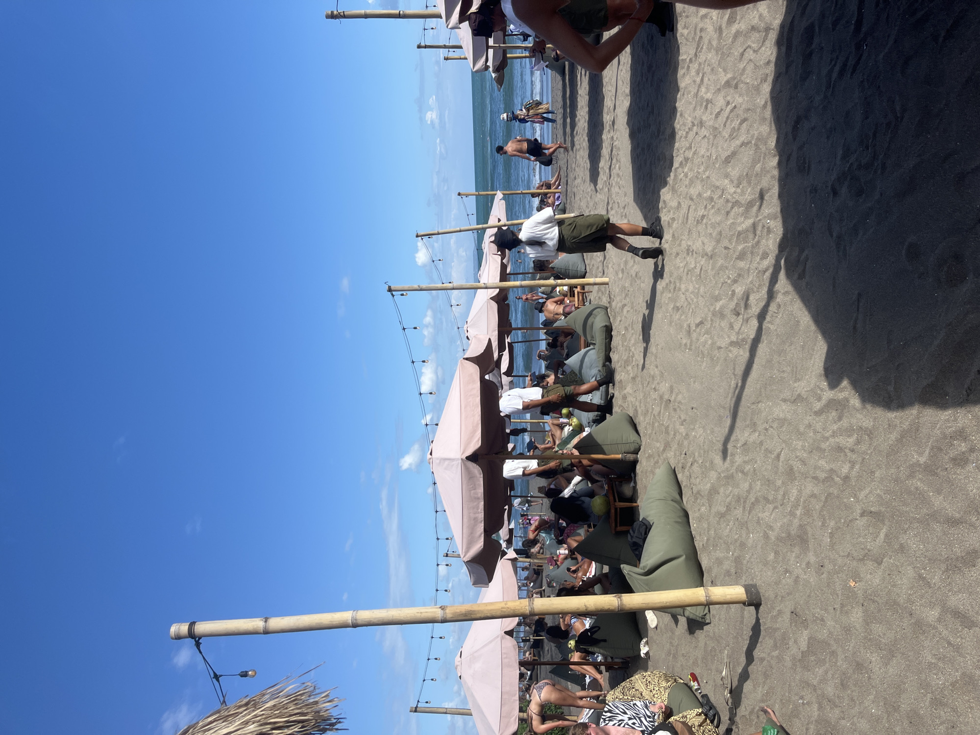 The image captures a beach scene in North Kuta, Indonesia, featuring a black sand beach lined with beach umbrellas and lounge areas. People are relaxing on beanbag chairs or walking along the shoreline. Several individuals are wading in the ocean. Bamboo poles support the umbrellas, and string lights are suspended from them. The sky is blue with a few clouds. A waiter in a white shirt and dark shorts is walking among the lounge areas.