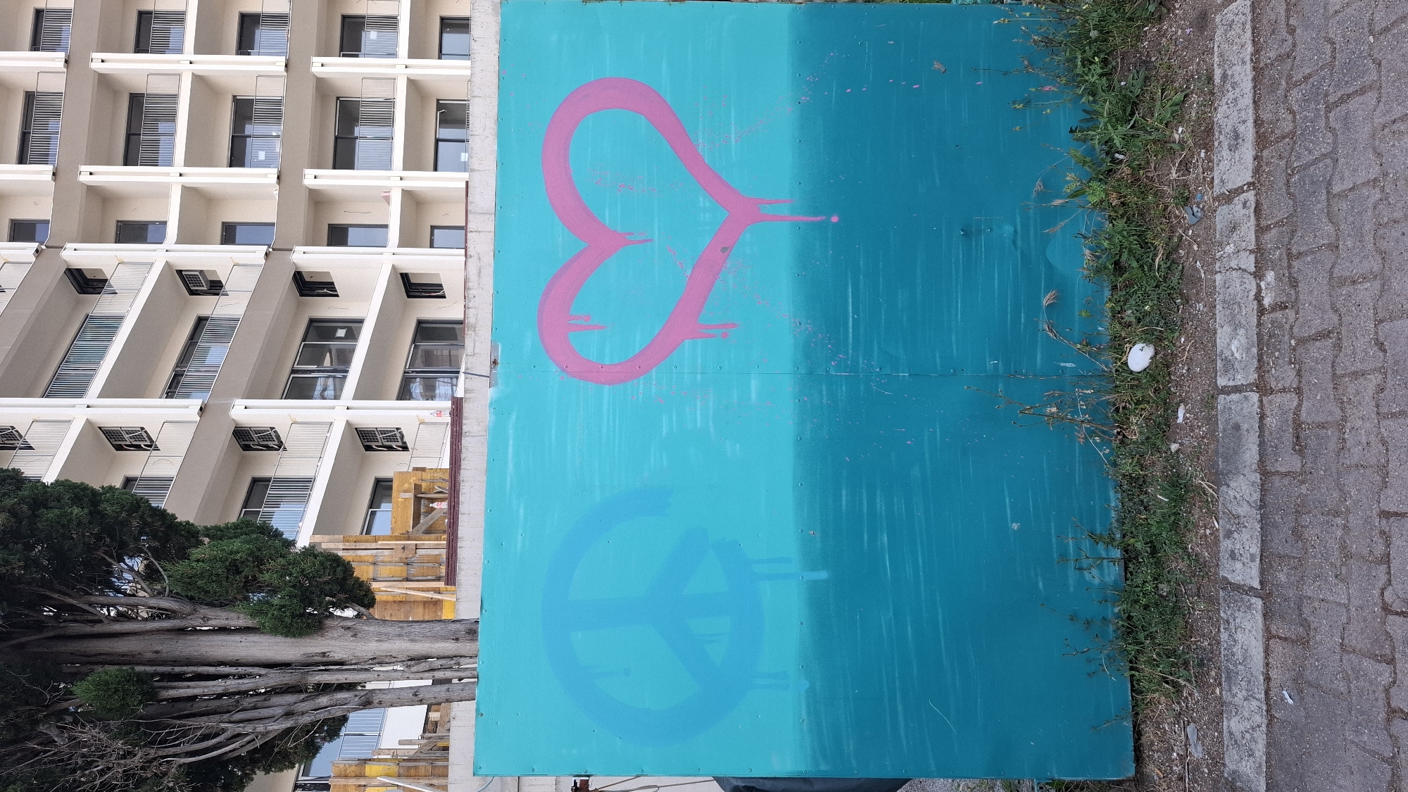 An exterior shot shows a two-paneled, light blue and turquoise painted barrier. On the top panel is a pink spray-painted heart-like symbol with arrow lines. Below, a light blue spray-painted symbol resembling a peace sign. To the left of the barrier is a building with multiple stacked apartments featuring square windows, along with a tree and construction materials. To the right, the barrier is bordered by green grass and a gray stone-tiled walkway. The photograph was taken in Bečići, Montenegro. No people are visible.