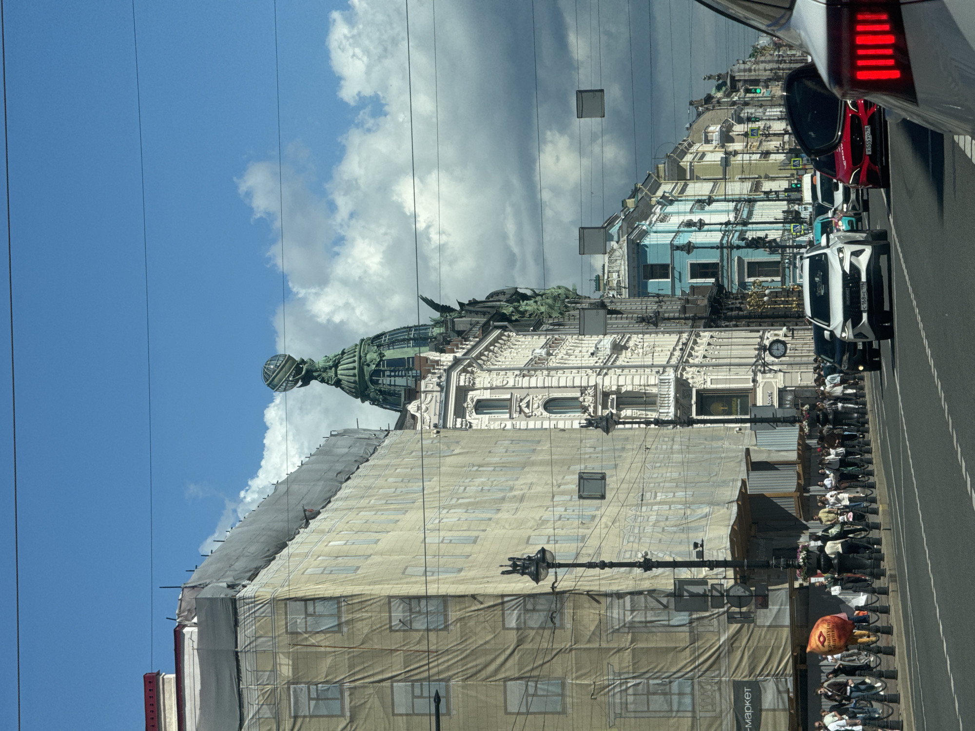 The image depicts a street scene in Saint Petersburg, Russia, likely on Nevsky Prospekt, given the architecture and the presence of the Singer House with its iconic globe on top. A large building on the left is covered in beige scaffolding and netting, suggesting ongoing construction or renovation. To its right, the Singer House stands with its green dome and distinctive globe.
The street is lined with various buildings showcasing different architectural styles and colors, including a bright blue facade. A sidewalk runs along the buildings, populated by a crowd of pedestrians. Many people are walking, some are standing in groups, and a few are engaged in conversations. A person in a bright orange, bulbous costume is visible near the sidewalk.
The road is busy with traffic. Several cars are moving along the street, including a white SUV, a red car, and other vehicles. The cars have license plates visible, indicating Russian registration. Traffic lights are visible, some showing green. Overhead, power lines stretch across the sky, a common feature in urban environments. The sky is a mix of bright blue and scattered clouds, suggesting a partly sunny day. Streetlights are also present along the sidewalk. The overall atmosphere is that of a vibrant, active urban environment in Saint Petersburg.