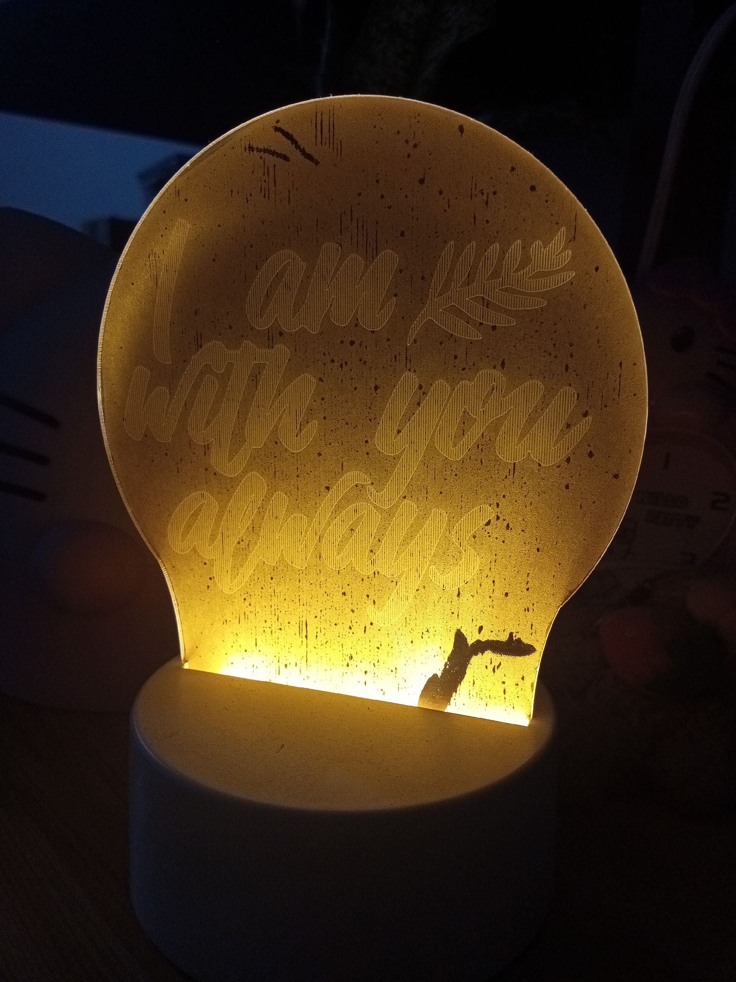 A close-up shot depicts a decorative night light illuminated in a dimly lit room. The night light consists of a white, cylindrical base supporting a clear acrylic or glass panel shaped like a bulb. The panel is laser-etched with the phrase "I am with you always," accompanied by a small floral or plant design. Light emanates from the base, illuminating the etched design, creating a warm, yellow glow. The panel has a speckled or textured appearance. The image is captured in a close perspective. It is impossible to verify that this image was taken in Davao City, Philippines, or any other specific location, without additional verifiable details.