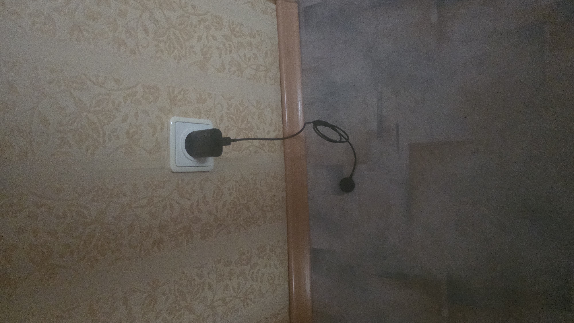 A black electrical adapter is plugged into a white wall socket on a wall with patterned wallpaper. A black cord extends from the adapter, looping onto the floor, where another black circular object is visible. The floor appears to be laminate or linoleum. A wooden baseboard runs along the bottom of the wall. The scene is likely indoors, possibly in a residential setting. There is no indication of location within Ukhta, Russia.