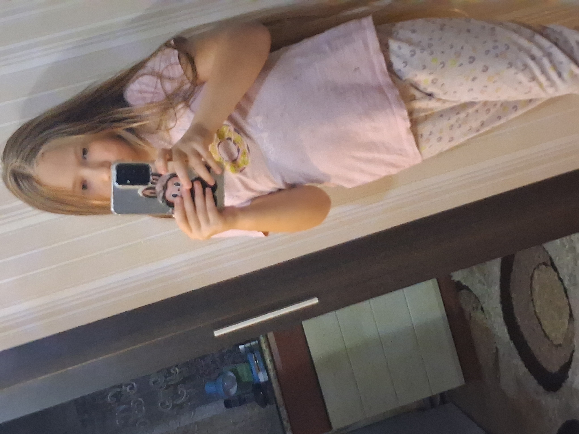 A young girl with long blonde hair takes a mirror selfie indoors. She is wearing a pink t-shirt with a donut graphic and patterned pajama bottoms. She is holding a smartphone with a cute case featuring a cartoon character. The background shows a wall with vertical stripes and a reflection of a closet with shelves and some objects, including a lamp. The floor has a patterned carpet. The location is not specified in the image or its metadata.