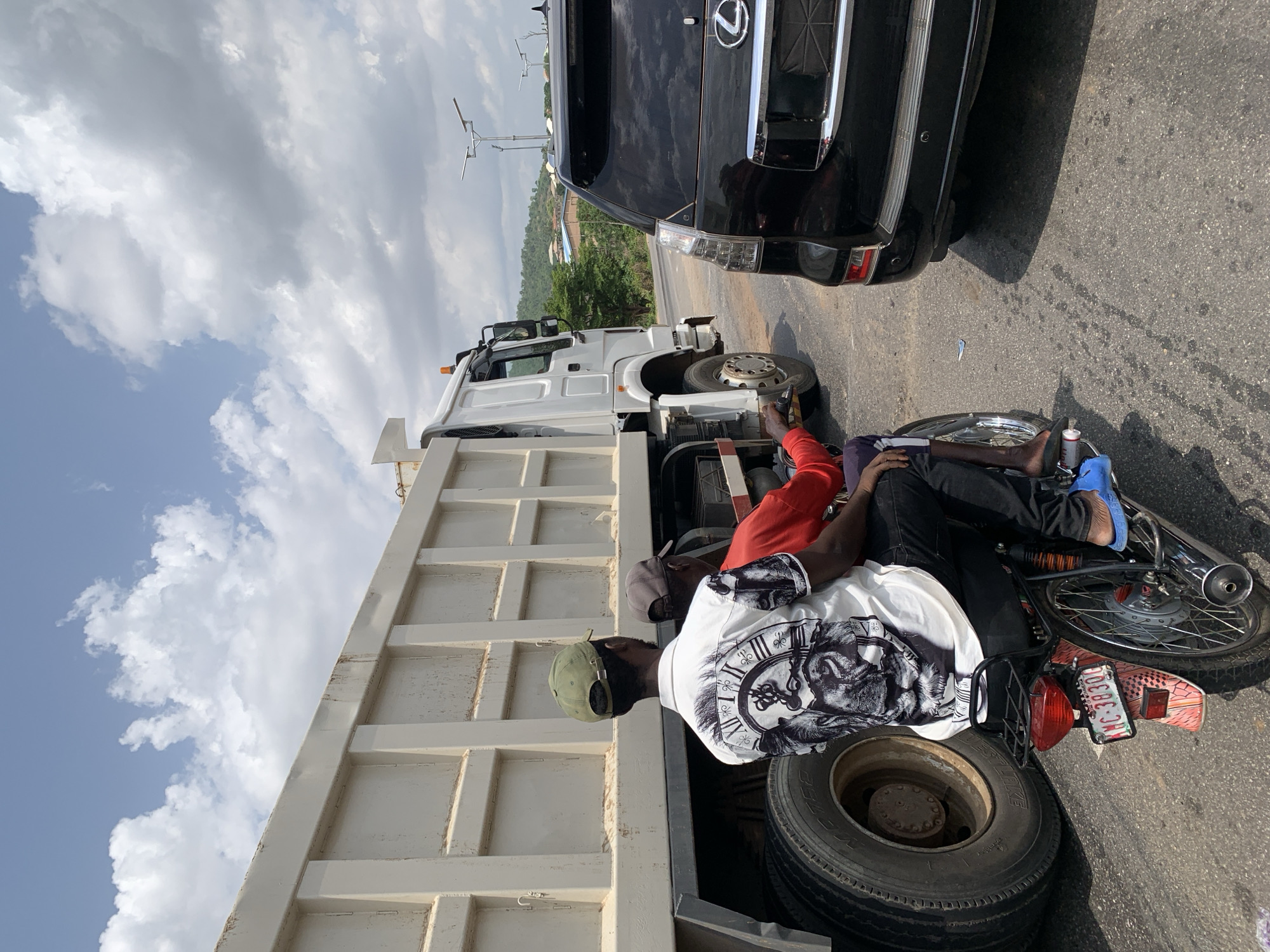 The scene is in Lokoja, Nigeria, on an asphalt road where a white dump truck is parked partially on the shoulder. Two men are near the truck. One man, wearing a white shirt with a black and white graphic of a face and clock, a green cap, and black pants, is sitting inside the truck bed. Another man, wearing a red shirt, is standing near the front of the truck, reaching towards the engine area. A black SUV is parked adjacent to the truck, encroaching into the roadway. A motorcycle is lying on its side near the truck, and its license plate is partially visible. The sky is partly cloudy.