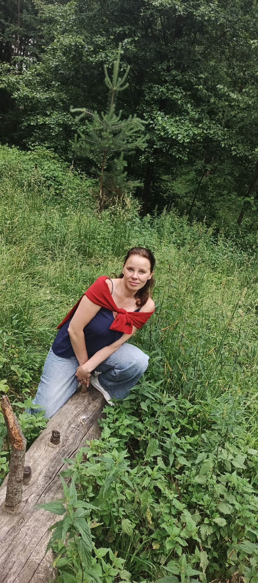 In the city of Chaikovsky, Russia, a woman is pictured in a natural setting. She is crouching on a fallen log amidst tall green grass and foliage. The woman is wearing a navy blue top with a red shawl draped over her shoulders, tied in the front. Her lower half is covered by light blue jeans and white sneakers. She has brown hair and is looking towards the camera. The background is dense with trees and various green plants.