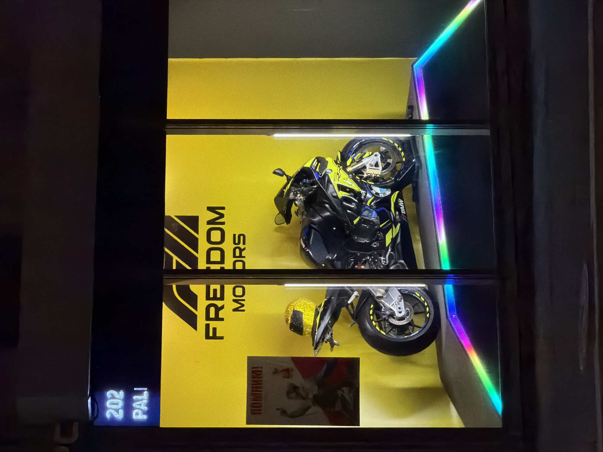 The image captures a storefront display, likely in Moscow, Russia, exhibiting a BMW motorcycle. The backdrop is a vibrant yellow wall emblazoned with the text "FREEDOM MOTORS" in bold, block letters. Below the motorcycle, a poster featuring a figure pointing is visible, with text in Russian. A matching yellow helmet is also part of the display. The storefront address, "202 PALI," is illuminated in blue at the bottom left corner. The display case is lined with colorful LED lights, adding to the modern aesthetic. The exterior setting appears to be at night, given the dark surroundings and the prominence of the illuminated display.