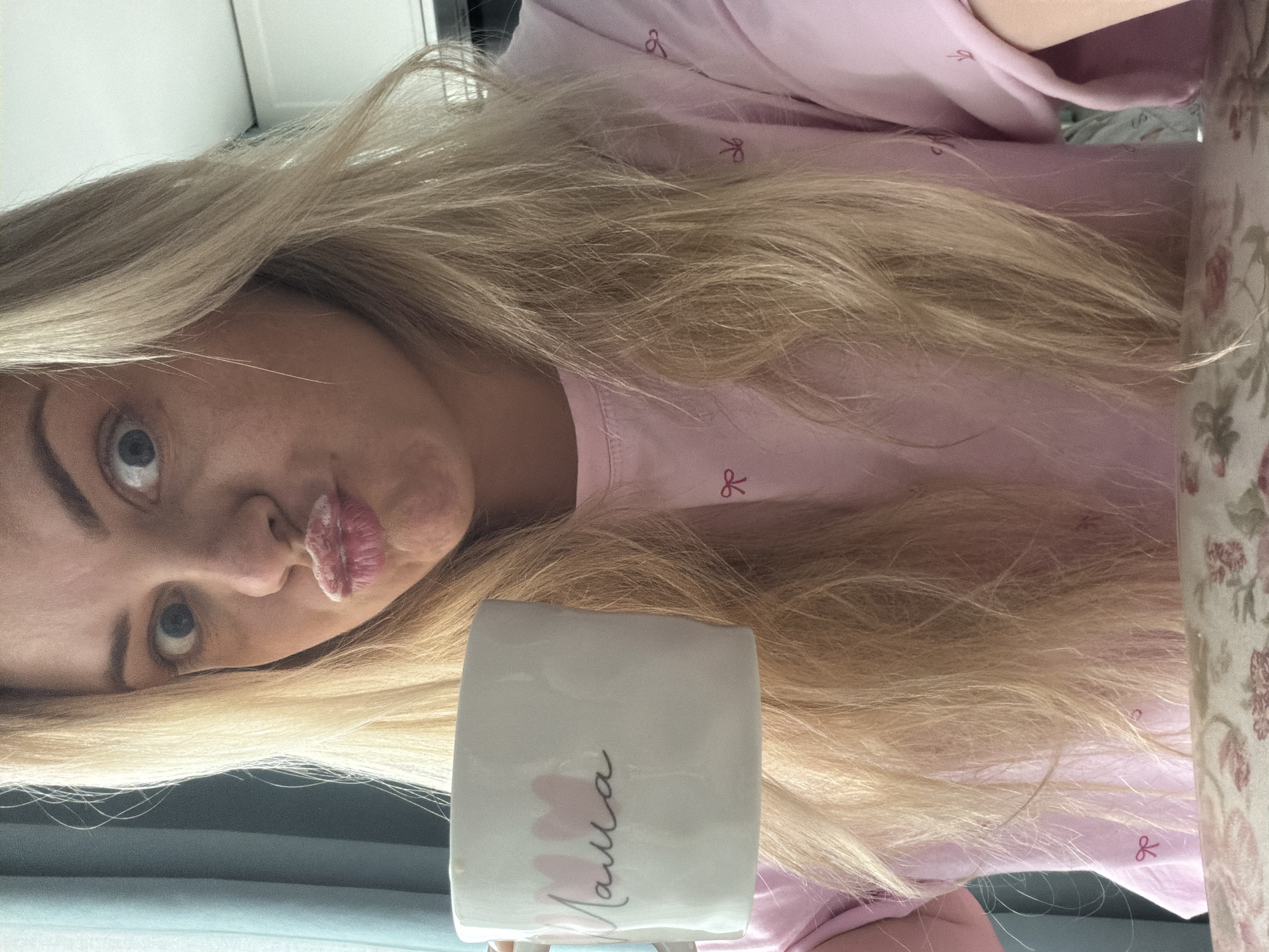 A playful selfie captures a young woman with long, straight blonde hair and fair skin. Her head is angled slightly, and she looks directly at the camera with wide, light-colored eyes. She is making a "kissing face" with her lips pursed, conveying a lighthearted and surprised expression.
She is wearing a light pink top decorated with small, darker pink bow patterns. In front of her, she holds a white ceramic mug, partially covered by her hair. The mug prominently displays the word "Мама" (Mama) written in a dark script font, accented with subtle pink brushstrokes.
The background suggests an indoor setting, possibly a home, with a glimpse of a floral-patterned fabric, likely a tablecloth or curtain, to her right. The lighting is bright, indicative of daytime. The overall scene is personal and casual. The context provided indicates the location is Votkinsk, Russia.