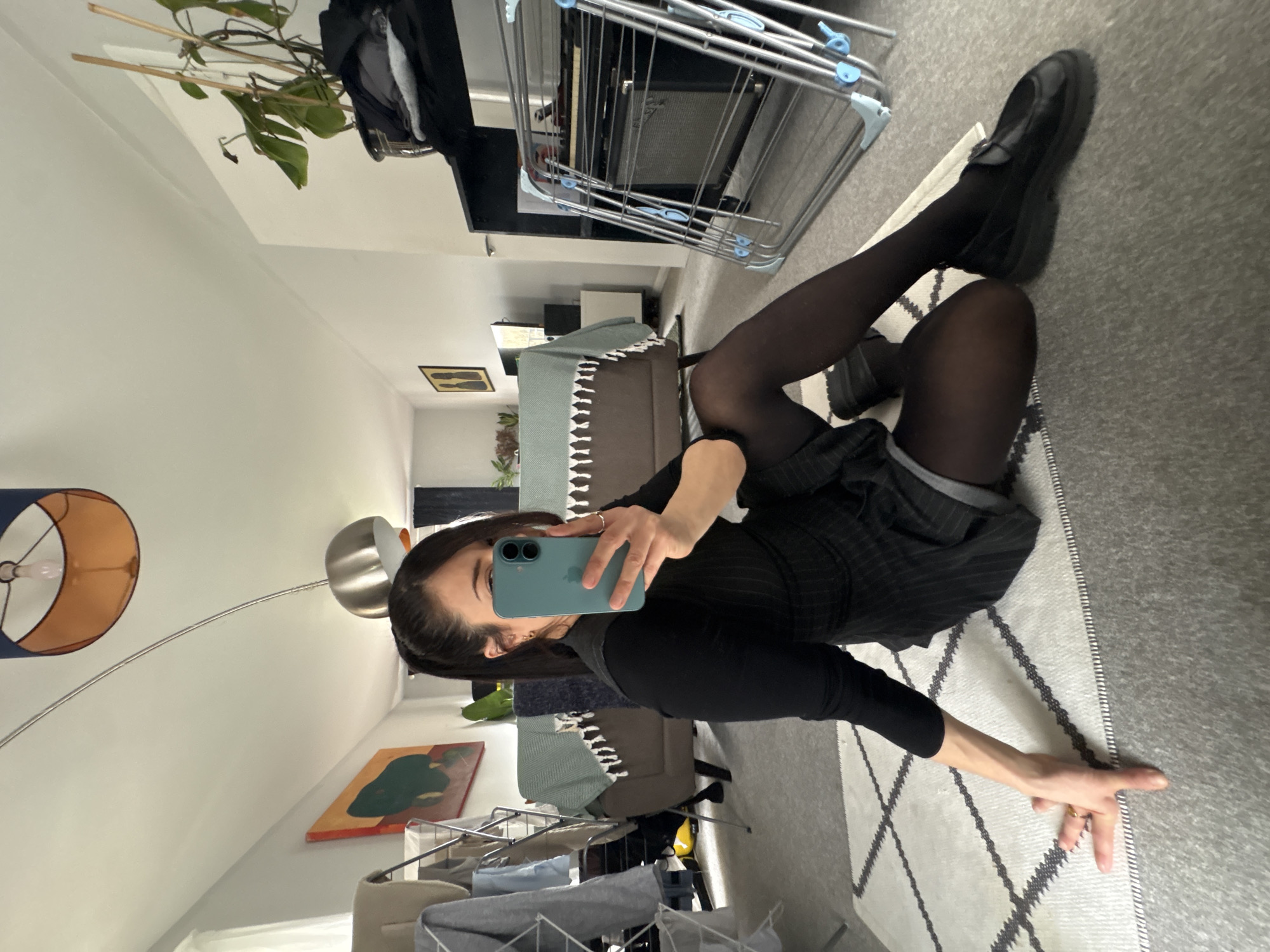 A woman sits on a rug indoors, taking a selfie. She wears a black long-sleeved top, a pinstripe skirt, black tights, and black shoes. She holds a turquoise phone up to take the photo. The rug has a geometric pattern of grey and white. Background items include a clothes drying rack, a lamp, furniture, and artwork on the wall. The location appears to be an apartment. There is no clear indication this is London, UK.