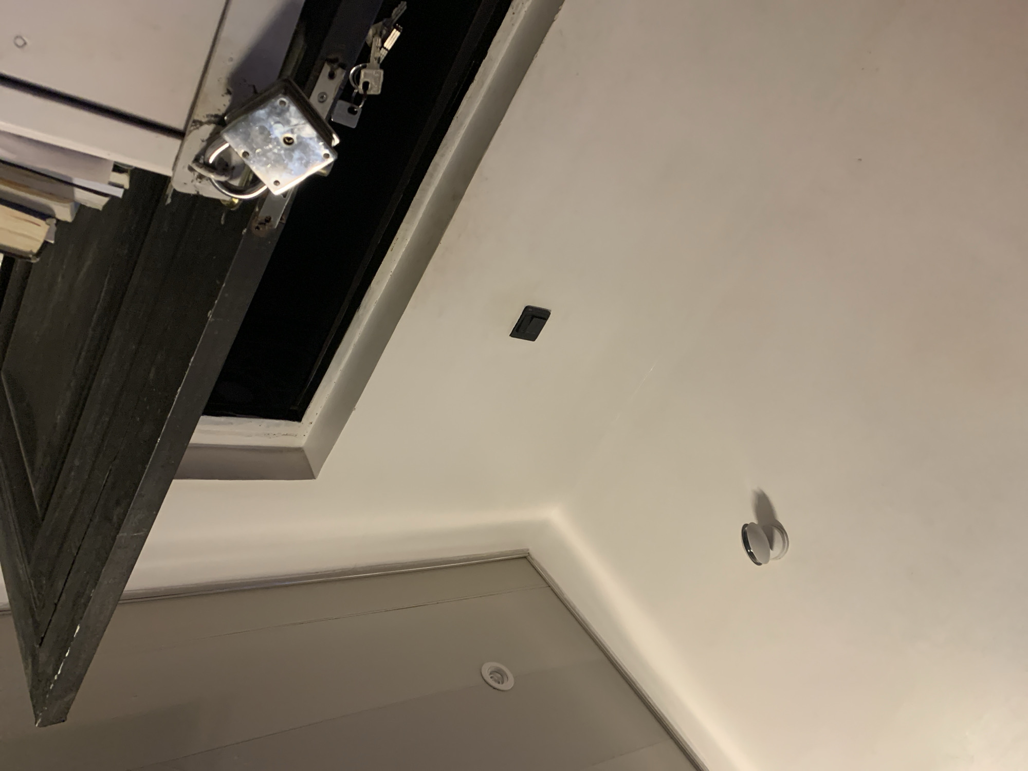 In an indoor scene of an Unknown, Unknown location, a white wall is visible. On the wall, near the top left, is a small white circular object with a black rim, possibly a security camera or sensor. Further down the wall, a small black rectangular object is mounted. In the lower right of the frame, a dark-colored door is ajar, revealing a dark interior. A silver padlock with keys attached is secured to the door frame. To the right of the padlock, a stack of books is visible. The ceiling is a light gray color with recessed lighting fixtures. The overall lighting suggests a dimly lit environment.