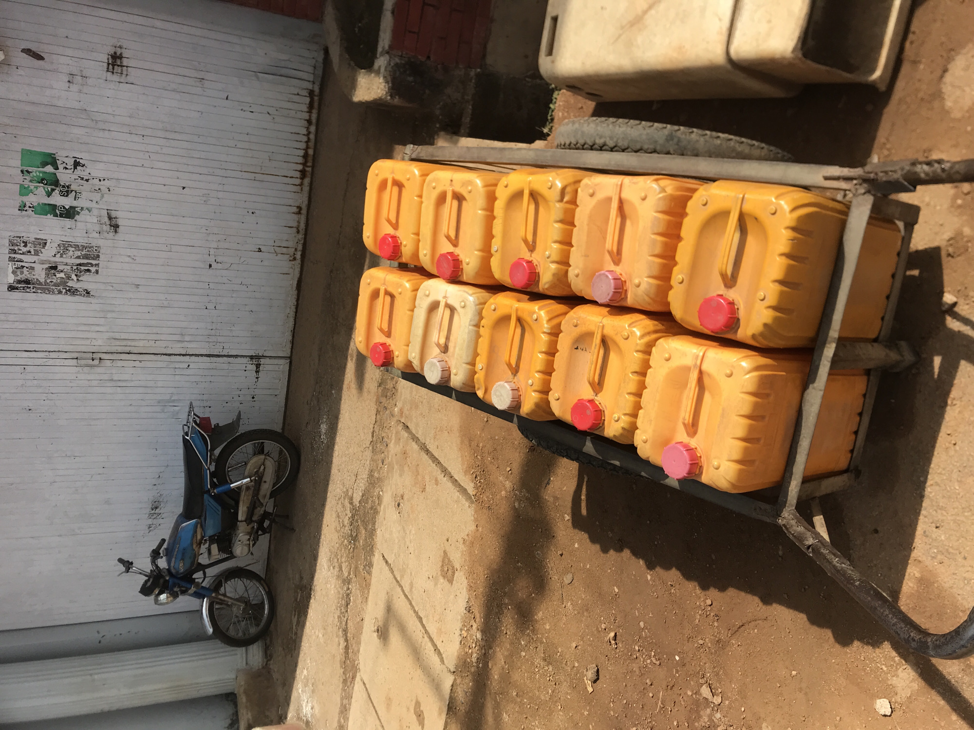 The image displays an outdoor scene in what appears to be an alleyway or side street in New Stadium, Nigeria, captured during bright daylight. The central focus is a metal cart or trolley, laden with eighteen orange plastic jerrycans stacked in two rows. Most of these jerrycans have red screw caps, while some feature lighter, possibly pink or white, caps. The cart is positioned on a ground composed of dry, reddish-brown dirt and some concrete slabs, and it casts a clear, distinct shadow.
To the left of the cart, a blue and black moped or small motorcycle is parked upright against a white, corrugated metal garage door. The door shows signs of wear and tear, with remnants of green and black markings or peeling posters visible. In the upper right background, partially obscured, is a large, off-white plastic container or tank, with a dark tire resting beneath it.
The strong shadows indicate a sunny, clear day, likely during mid-morning or afternoon. No individuals are visible in the frame. The presence and arrangement of the jerrycans on the cart suggest they are either being transported or are part of an informal vending operation, commonly used for fuel or water in the region. No legible text is visible on any of the objects or surfaces.