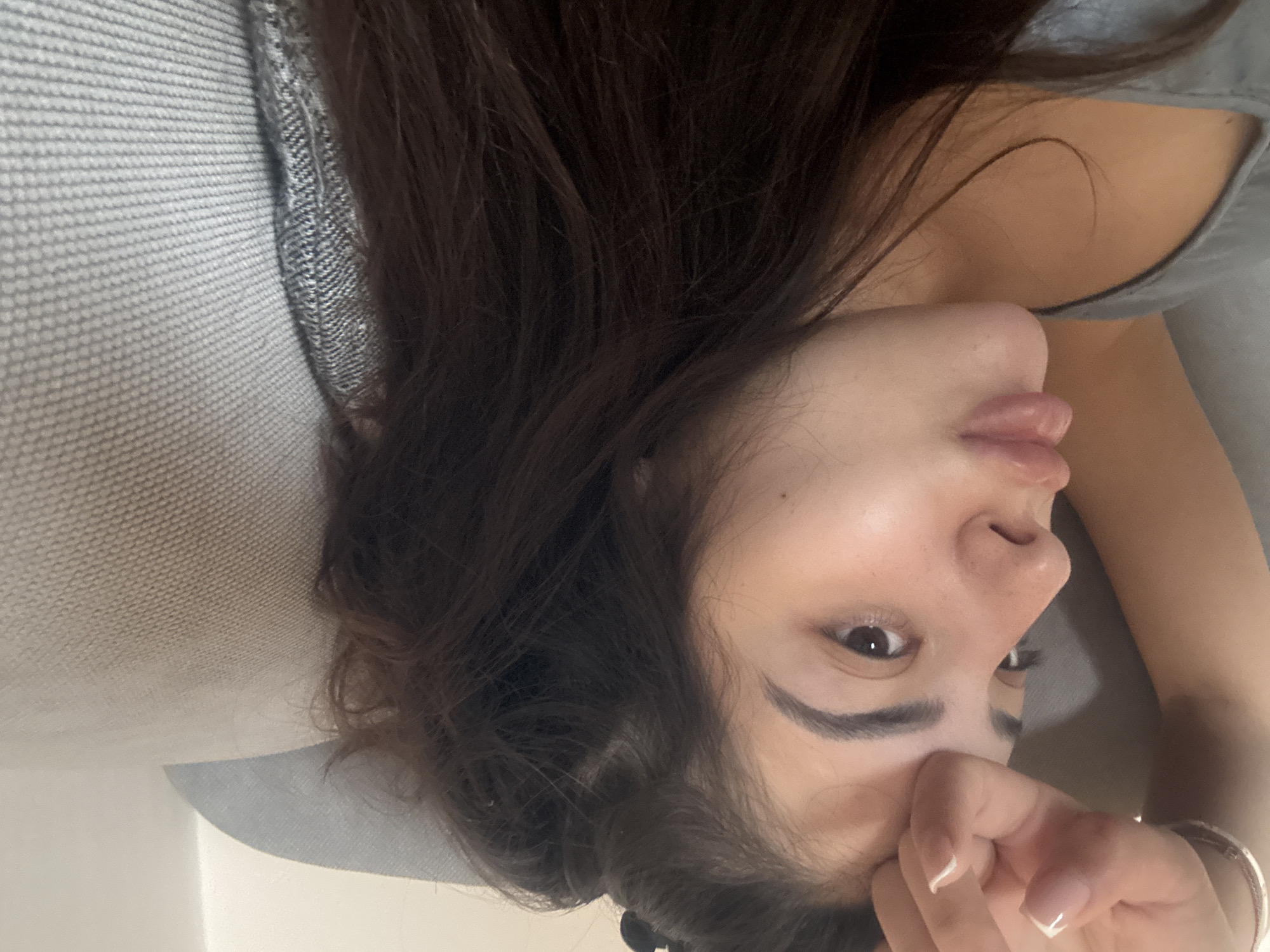 The image presents a close-up, intimate portrait of a person, likely female, captured from an overhead perspective and rotated 90 degrees clockwise. The individual has long, dark brown hair that partially obscures the face and drapes across the shoulder. They are lying on a light grey, subtly textured fabric surface, suggesting a sofa or bed in an indoor setting.
Visible features of the face include a dark eye with a well-defined dark eyebrow, part of the nose, and slightly parted, naturally glossy lips. The expression appears relaxed and somewhat pensive, with the gaze directed subtly away from the camera. One hand is gently resting near the chin/mouth area, revealing fingernails with a clean French manicure style. A thin, light-colored bangle or bracelet adorns the wrist of this hand.
The person is wearing a casual light grey garment, possibly a tank top or camisole, with a textured strap visible near the shoulder. The lighting is soft and diffuse, indicating an indoor environment, likely during daytime, though no specific time of day or weather conditions are discernible. There is no visible text in the image. The scene conveys a moment of quiet repose or casual posing for a photograph.