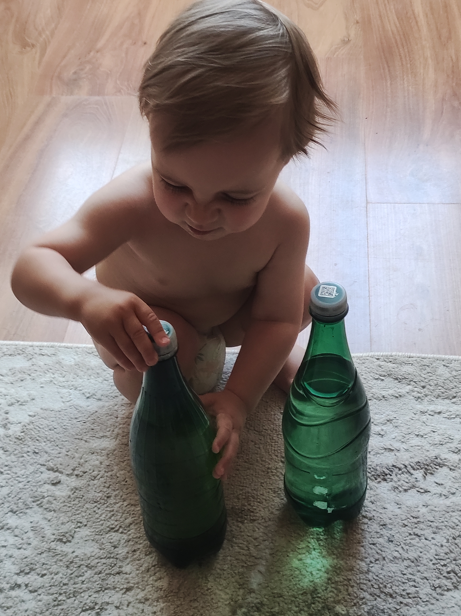 A child is playing with two green glass bottles on a light-colored, textured rug. The child, who appears to be fair-skinned with light brown hair, is unclothed. One hand rests on the top of the bottle on the left. The bottle on the right has a QR code on the lid. The location is indicated as Chaikovsky, Russia, although there is no visible evidence in the image to support this. The background shows a light-colored wooden floor.