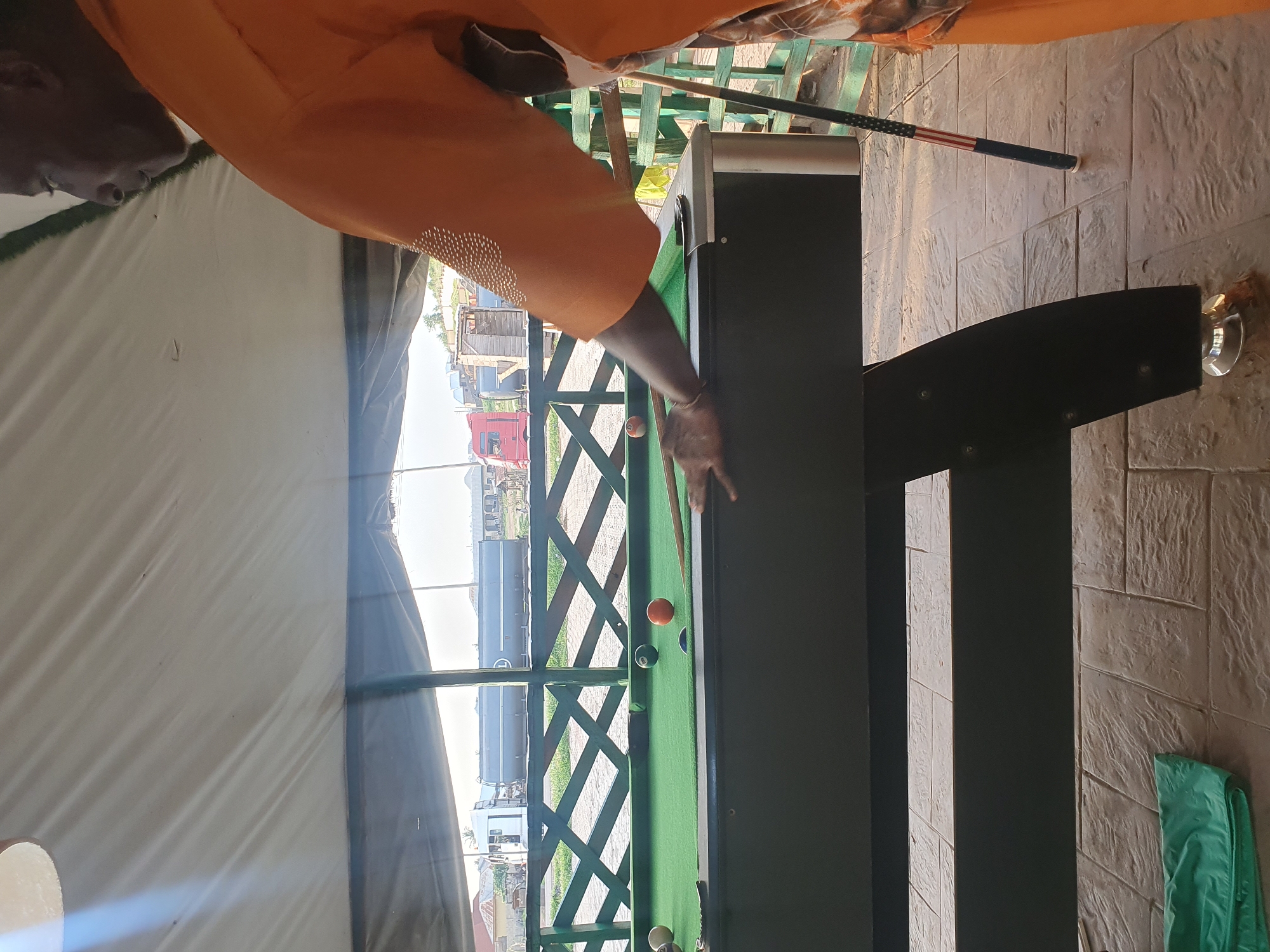 The image shows a person playing pool at an outdoor establishment. The person, wearing an orange shirt, is aiming a cue stick at a pool ball on a green felt table. The scene appears to be set in Lokoja, Nigeria, given the context provided. The background reveals a partially visible white tent or canopy, suggesting an open-air setting. Beyond the tent, there are structures that look like shipping containers or buildings, and some vegetation. The ground is paved with textured tiles. A pool cue with an American flag design is visible leaning against a green wooden structure in the background. The lighting suggests it might be daytime, possibly late afternoon, with some shadows present. The overall mood seems relaxed and recreational.
