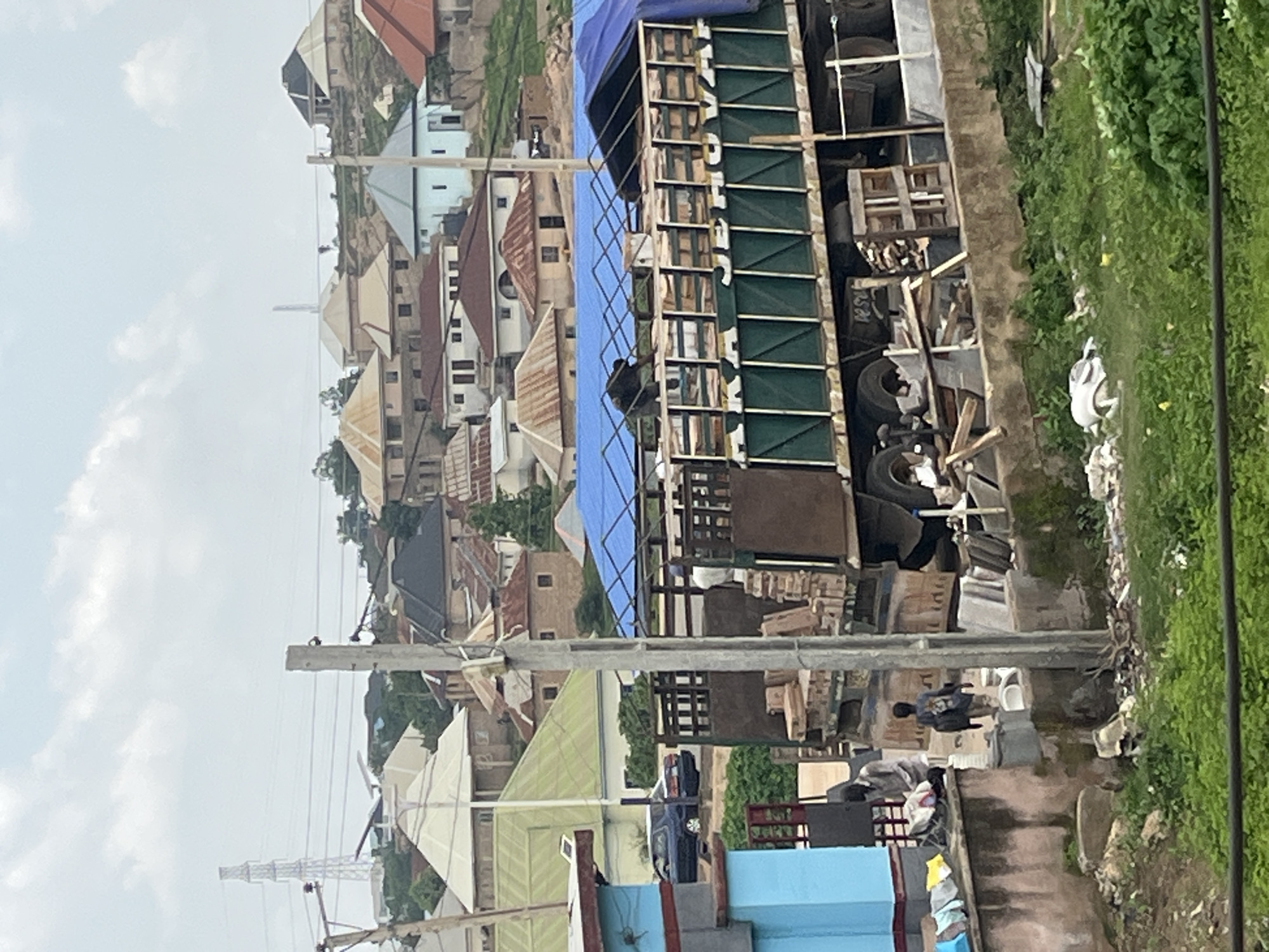 This image depicts an outdoor scene in Gura Top, Nigeria. The perspective is from a high vantage point, looking down at a densely populated area with numerous buildings characterized by their varied roofing styles, including red, silver, and brown. The architecture suggests a mix of residential and possibly commercial structures.
In the foreground, a large truck with a blue tarp covering its bed is prominently featured. The side of the truck has green slatted panels and appears to be carrying various materials, including what look like tires, wooden planks, and other assorted items. A person is visible near the front of the truck, appearing to be interacting with the cargo.
Further into the scene, there are more buildings, some with visible inhabitants and activities. A dark-colored vehicle is parked on a pathway. In the middle ground, there are more structures and a pathway or road, with some debris visible along the side. A person can be seen in this area, near what appears to be an outdoor toilet.
The background shows more of the settlement extending up a gentle slope. A tall transmission tower is visible in the distance, and electrical lines crisscross the frame, indicating the presence of infrastructure. The sky is overcast with clouds, suggesting a potentially humid or rainy weather condition. The overall impression is one of a busy, developing urban or semi-urban environment, with a sense of everyday life and commerce. Some of the text visible on the truck's side appears to be "JETOVA", though it's partially obscured.