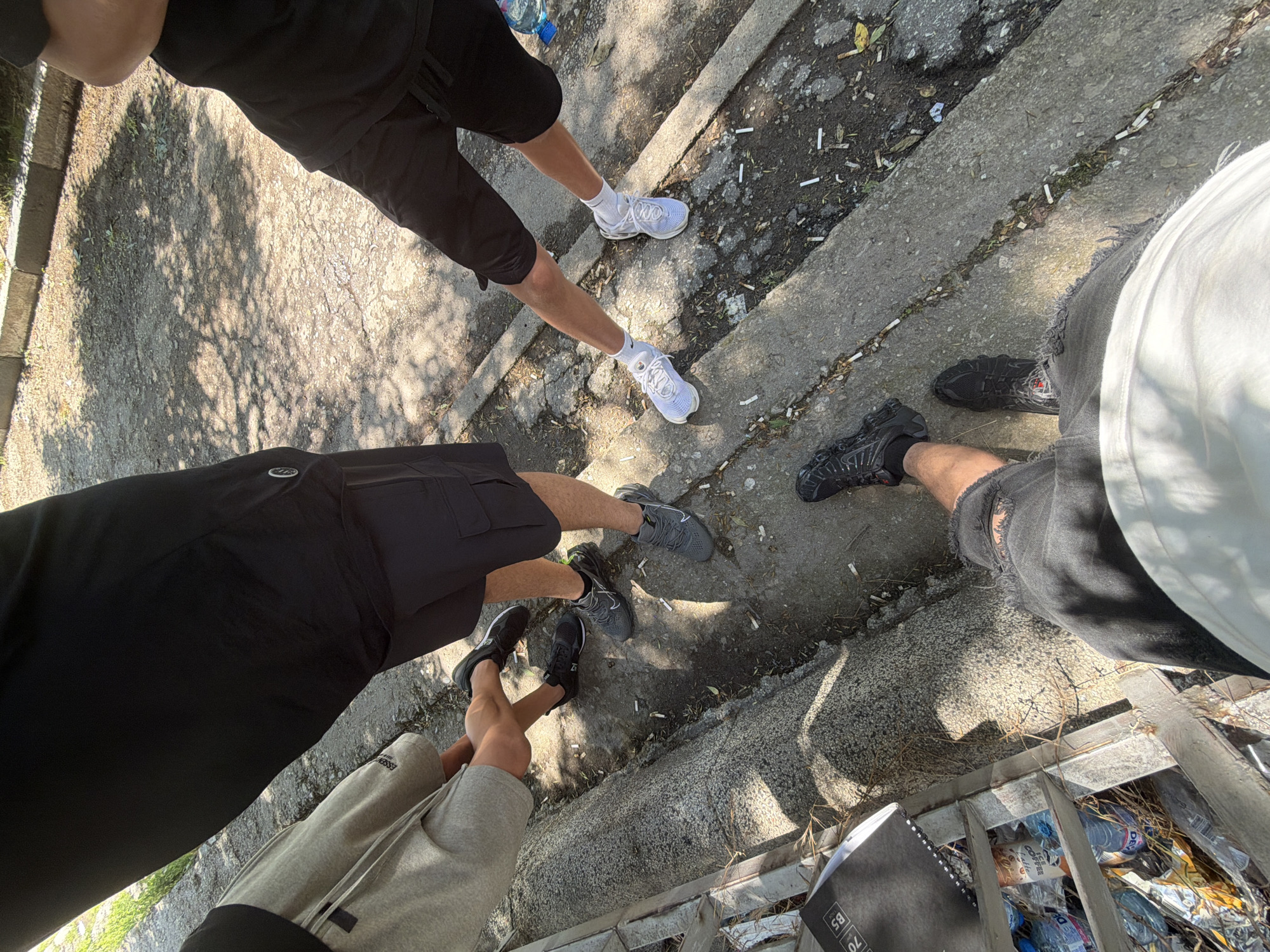 The image captures a low-angle view of four individuals' legs and feet, positioned outdoors on a sun-dappled, uneven paved surface that appears to be a public space within the city of Plovdiv, Bulgaria. The ground is a mix of asphalt and concrete, with visible cigarette butts scattered throughout.
The people are casually dressed in shorts and athletic shoes. From left to right, the first person's legs are clad in grey shorts and black sneakers. The second person wears black shorts and grey sneakers. The third individual sports black shorts and white sneakers with white socks. The fourth person, closest to the viewer, is seen from the waist down, wearing black distressed shorts and black athletic shoes. One of the individuals on the right appears to be holding a clear plastic bottle of water.
The background reveals a textured concrete barrier or ledge, a patch of greenery, and what appears to be a pile of discarded items, including plastic bottles, in a metal bin or crate on the left side of the frame, suggesting a less maintained area of the city. The sunlight creates strong shadows, indicating it is daytime. The overall scene depicts a casual, informal gathering in an urban outdoor environment.