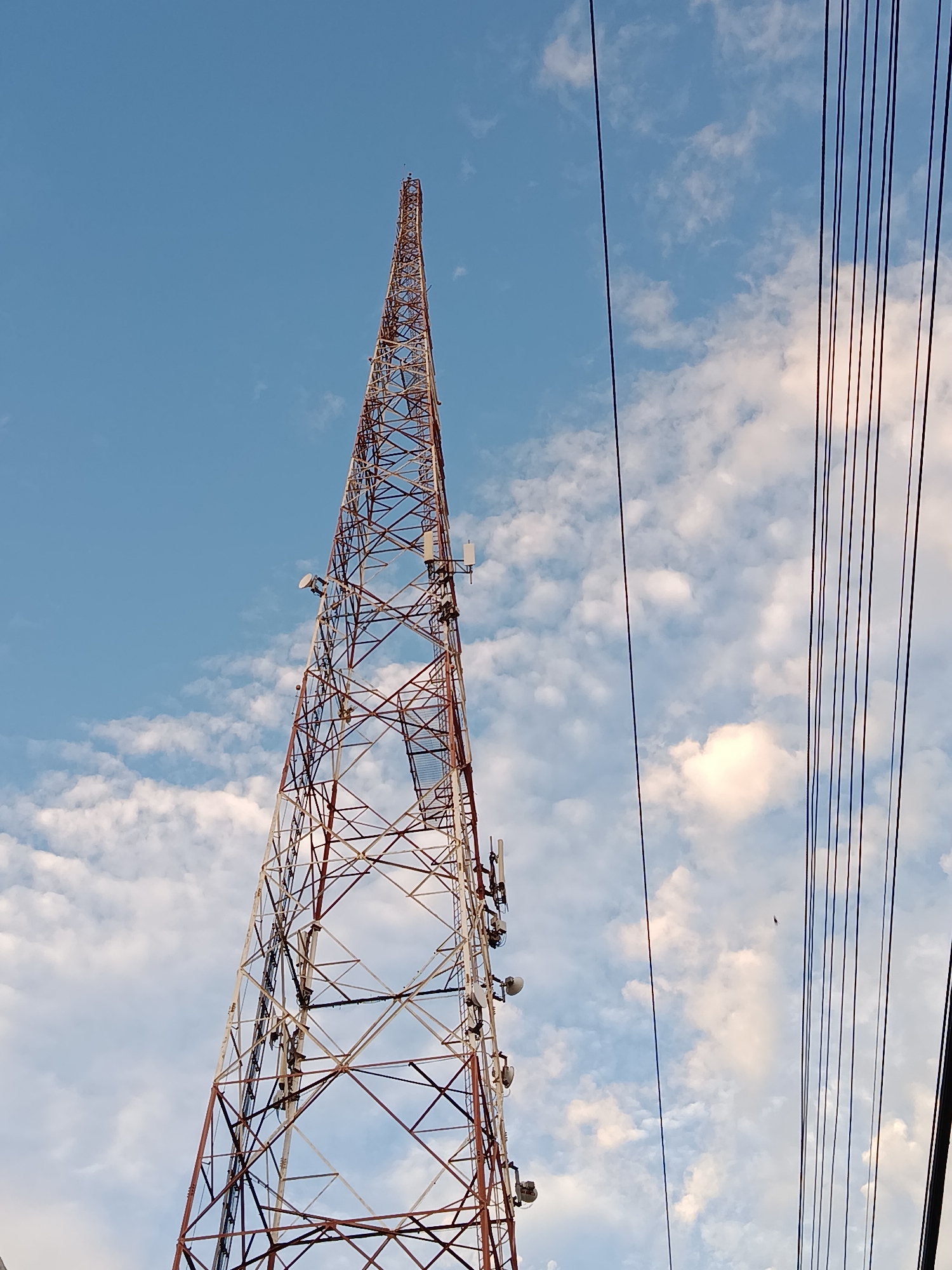 The image shows a tall, red and white lattice tower, likely a telecommunications tower, reaching into a blue sky filled with scattered white clouds. Multiple antennas and equipment are mounted on the tower. To the right of the tower, a set of parallel black wires runs vertically through the frame. The perspective is from below, looking upwards. The scene appears to be an outdoor urban or semi-urban environment.