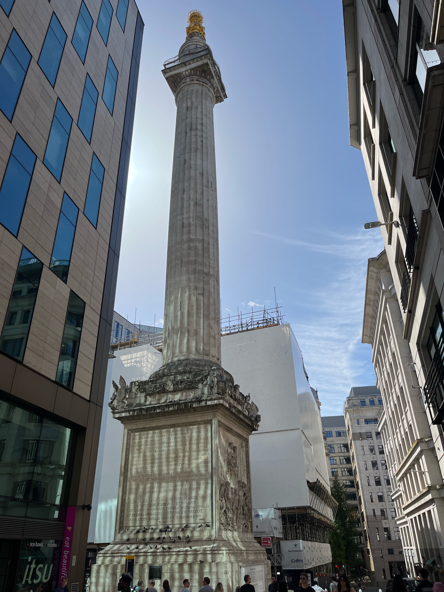 The image captures a view of The Monument, a tall Doric column situated in the City of London, United Kingdom. The Monument stands prominently in the center of the frame, surrounded by modern and classic architecture. The column itself is made of stone and features intricate carvings on its base. Above the base, the column is fluted and topped with a golden orb. The sky is a clear blue with a few wispy clouds.
To the left of The Monument, a contemporary building with a facade of horizontal beige panels and regularly spaced blue-tinted windows rises. Its reflection is visible in some of these windows. On the right, a light-colored classical building with many windows and dark shutters is visible, along with other buildings in the background.
In the middle ground, behind The Monument, a large building is under construction or renovation, covered in white sheeting and surrounded by scaffolding. Signs with logos like "Collins" and "North Standard" are visible on the sheeting.
At the base of The Monument, several people are gathered, some walking and others standing. They are dressed in casual summer clothing. There are also a number of pigeons on the ground around the monument. The scene suggests a busy urban environment on a sunny day.