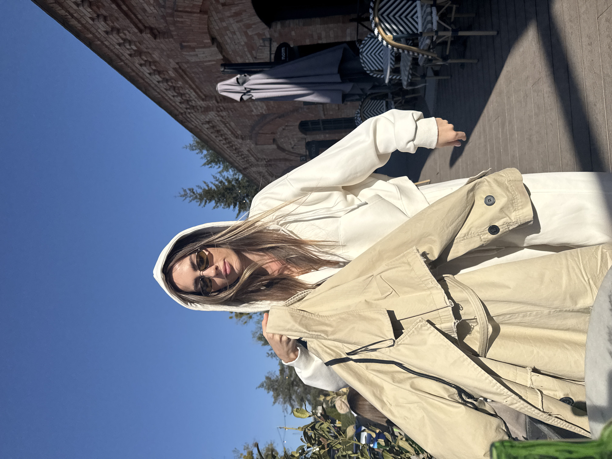 An outdoor portrait captured on a bright, sunny day features a young woman looking directly at the camera. She has long, light brown hair and is wearing a white hooded sweatshirt and amber-tinted sunglasses. In her arms, she holds a light beige trench coat.
The scene is set in what appears to be an urban outdoor cafe or pedestrian area. Behind the woman, there is a distinct brick building with architectural details. To her right, elements of an outdoor cafe are visible, including stylish black and white patterned chairs, tables, and a closed grey umbrella. The ground is made of wooden decking, and strong, defined shadows suggest direct sunlight, indicating mid-day or early afternoon. A clear blue sky dominates the upper portion of the frame, and some green foliage from trees is seen on the lower left. The partial word "nom" is visible on a sign or object near the cafe furniture. The overall impression is one of casual comfort in a sunny, city environment, fitting for a location like Warsaw, Poland.