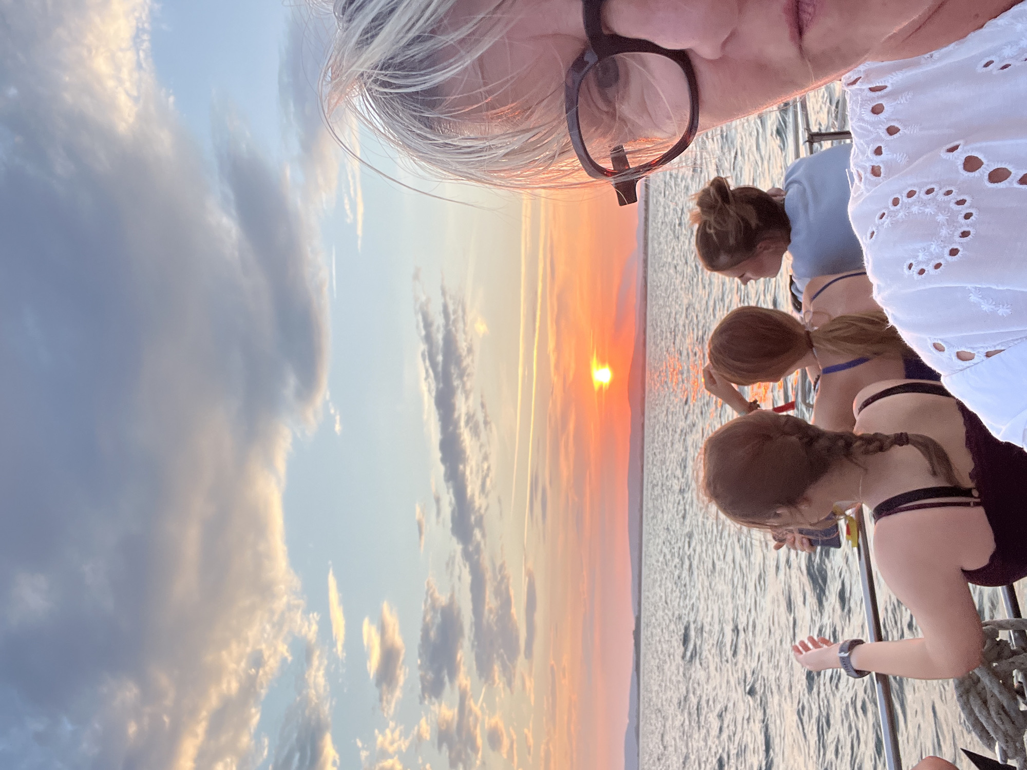 A medium shot shows four people on a boat at sunset, with the sun setting over a body of water and a distant coastline. The foreground is dominated by a woman's face, looking towards the camera and wearing glasses and a white embroidered top. Behind her, three other individuals are visible. The person on the far left has their back to the camera, wearing a dark top, and is looking out at the sunset. To their right, another person is also looking away, wearing a blue top. The third person, on the far right, is partially visible and appears to be holding a drink. The sky is filled with dramatic clouds, tinged with the warm colors of the sunset. The water is calm, with gentle ripples reflecting the sky's hues. The scene captures a serene moment during a boat trip, likely in Palavas-les-Flots, France, given the characteristic coastal landscape and sunset over the Mediterranean Sea.