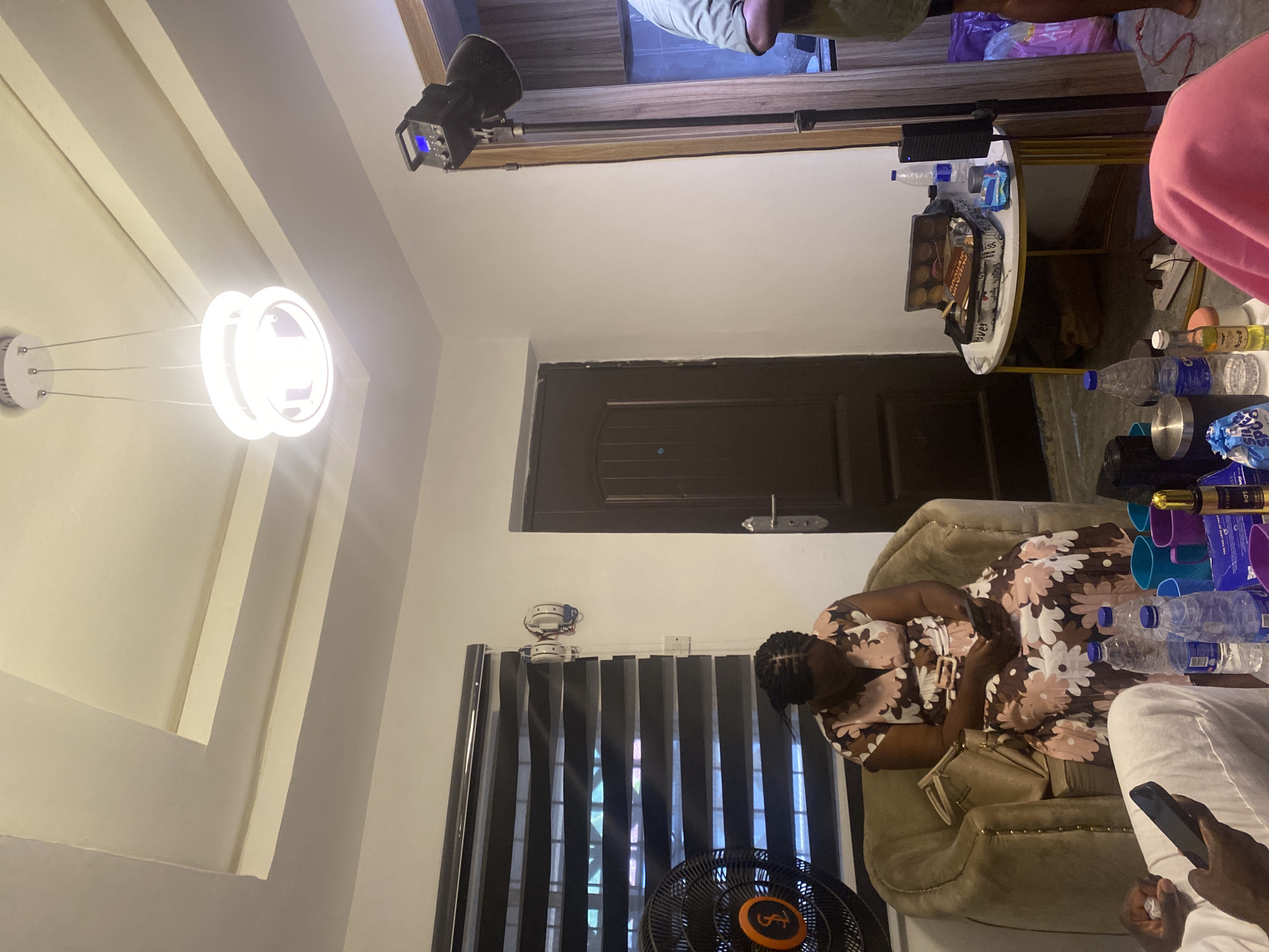 A cluttered interior in Ado-Ekiti, Nigeria. A woman in a floral dress sits in an armchair, likely using a phone. A ceiling fan stands near a window with blinds. A chandelier hangs from the ceiling. Bottles and other items clutter a table. Other people are present. A door is in the background. Lighting equipment is present.