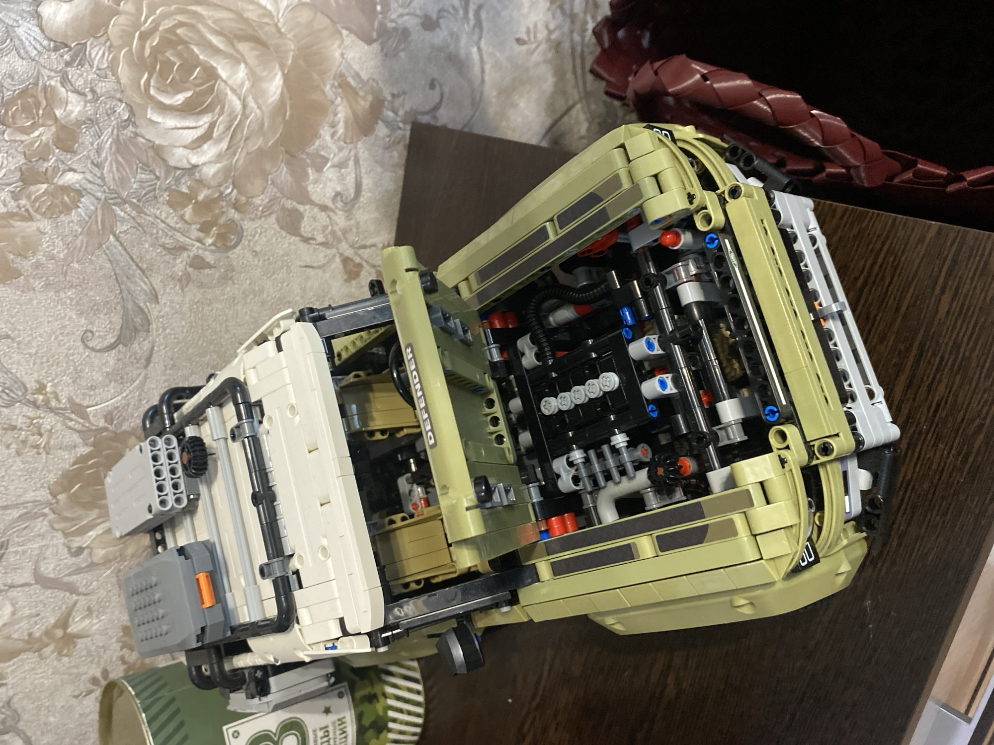 An unassembled Lego Technic Land Rover Defender is displayed on a dark wooden table, set against a wall with floral wallpaper. The model is primarily tan and white, with exposed mechanical components, including the engine, visible under the lifted hood and bonnet. Two gray Lego accessories, one resembling a storage box and the other a rocket launcher, are positioned on the roof rack. A cylindrical green object and a fragment of a green and white label with Cyrillic text are partially visible to the left of the model. To the right of the model, a segment of a dark red, woven object is present. The lighting suggests an indoor setting. The wallpaper features a textured, cream-colored background with large, embossed roses in shades of pink and cream. The overall scene is static, with no people or overt actions depicted.