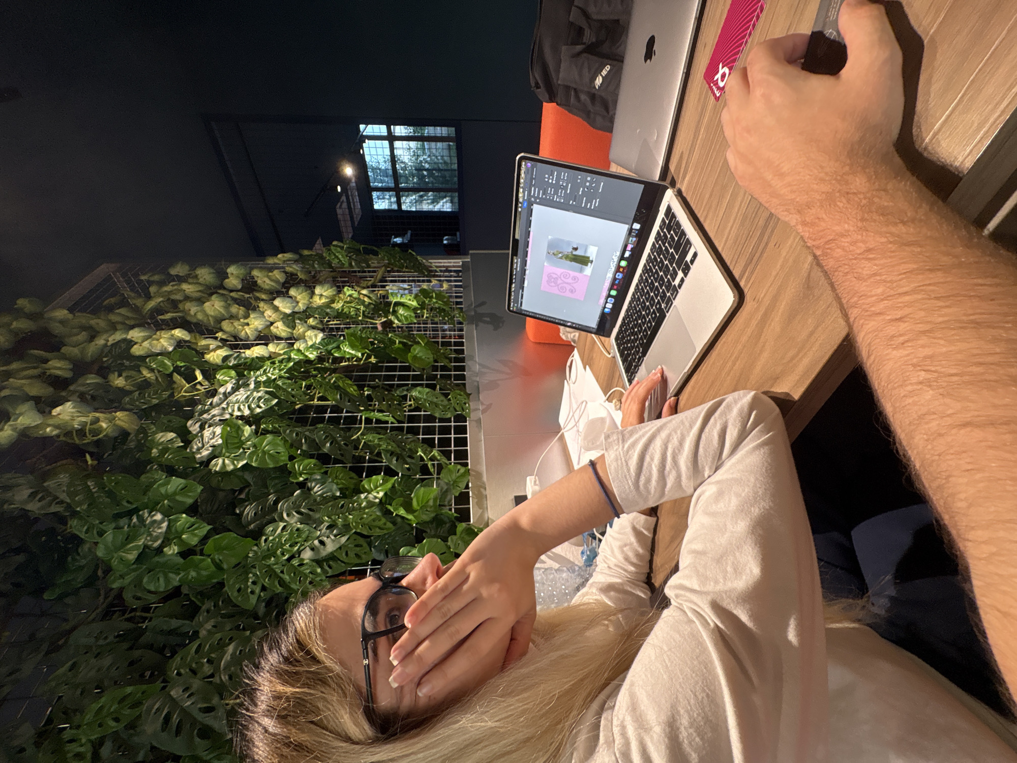 The image depicts an indoor scene in what appears to be a modern co-working space or office in Novate Milanese, Italy. Two individuals are seated at a light brown wooden desk.
On the left, a blonde-haired female with fair skin is seen in profile, wearing black-rimmed glasses with a visible logo on the temple (Chanel). She is wearing a light-colored long-sleeve top. Her left hand supports her chin and cheek, with her elbow resting on the desk. Her right hand is positioned over the trackpad of an open silver MacBook Air laptop. A thin, dark blue bracelet is visible on her right wrist. Her fingernails are manicured. The laptop screen displays graphic design software, likely Adobe Photoshop, showing an image of a person in a green outfit against a pink background, with various toolbars and layers visible. A pair of dark blue trousers are visible beneath the desk.
To the right, partially obscured, is the hairy forearm and hand of a male individual, also fair-skinned, resting on the desk. His hand holds a dark, rectangular object, possibly a computer mouse. Adjacent to his hand on the desk is a pink card with a white alpha symbol ("α").
Behind the open laptop, a closed silver MacBook is present, and a black fabric bag with a white "IED" logo is visible behind it.
The background features a prominent vertical garden wall composed of various lush green plants, including *Monstera Adansonii* (Swiss cheese plant) and other leafy vines, mounted on a metal grid structure. This plant wall is well-lit from above. To the right of the plant wall, the room transitions to a dark blue or grey wall. A rectangular window with a grid pattern is visible in this wall, revealing a blurred view of trees and ambient light from outside, suggesting daytime. An orange segment, possibly part of a bench or seating, is visible behind the laptops. White cables are seen on the desk surface near the left laptop.